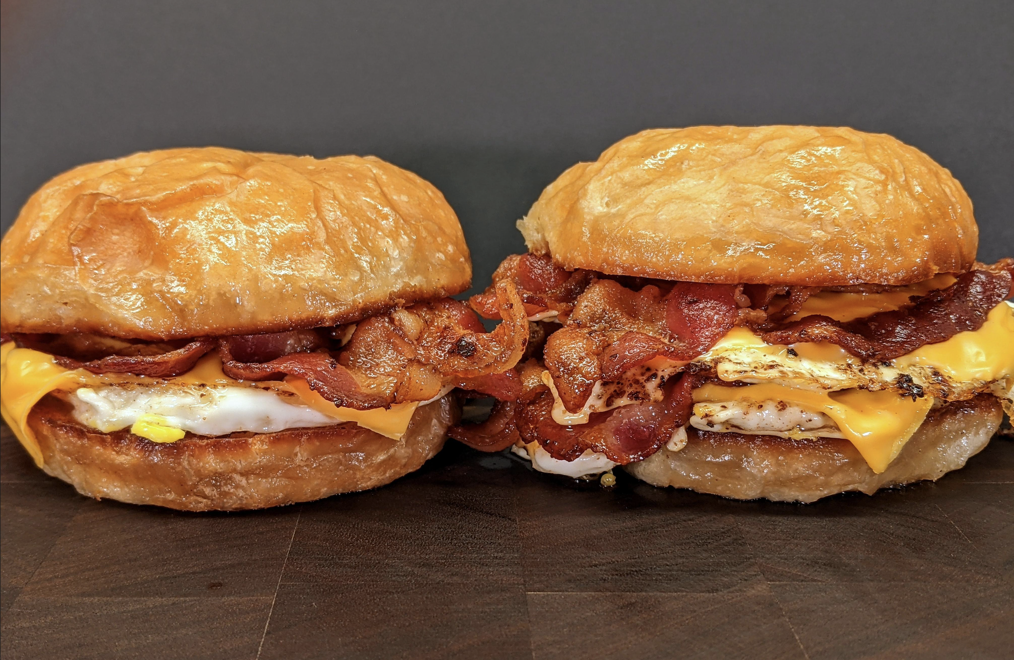 Two breakfast sandwiches with fried eggs, crispy bacon, and melted cheddar cheese on shiny, golden croissant buns, placed side by side on a dark wooden surface with a plain dark background.