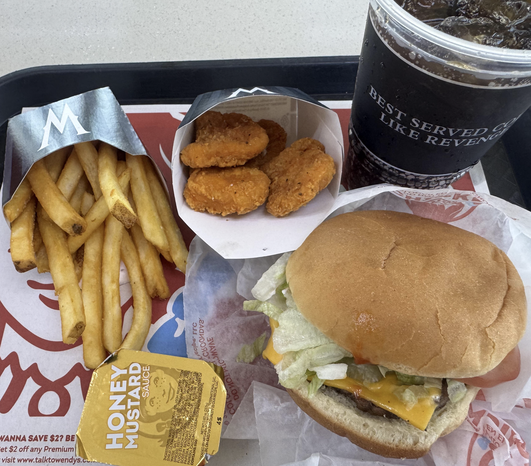 Fast food meal with fries, a cheeseburger with lettuce, chicken nuggets, honey mustard sauce, and a large soda on a tray.