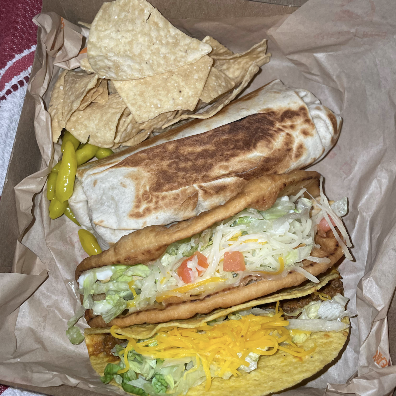 A takeout box with tortilla chips, pickled peppers, a grilled burrito, a fry bread taco topped with lettuce and cheese, and a crispy taco with shredded cheddar, lettuce, and tomato, all on parchment paper.