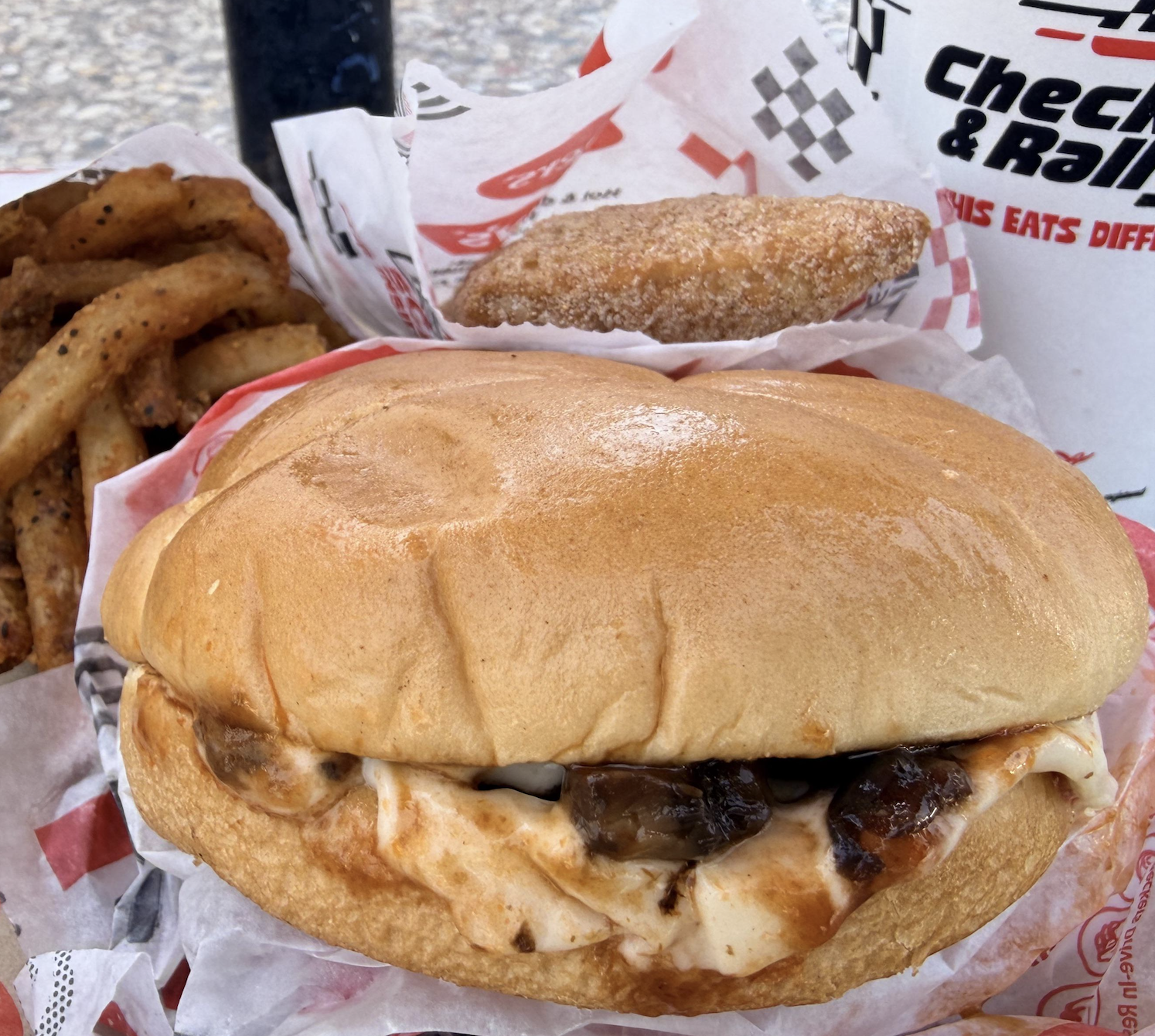 A sandwich with melted cheese and onions, seasoned fries, a cinnamon sugar donut, and a drink cup from Checkers & Rally’s, all on a tray.