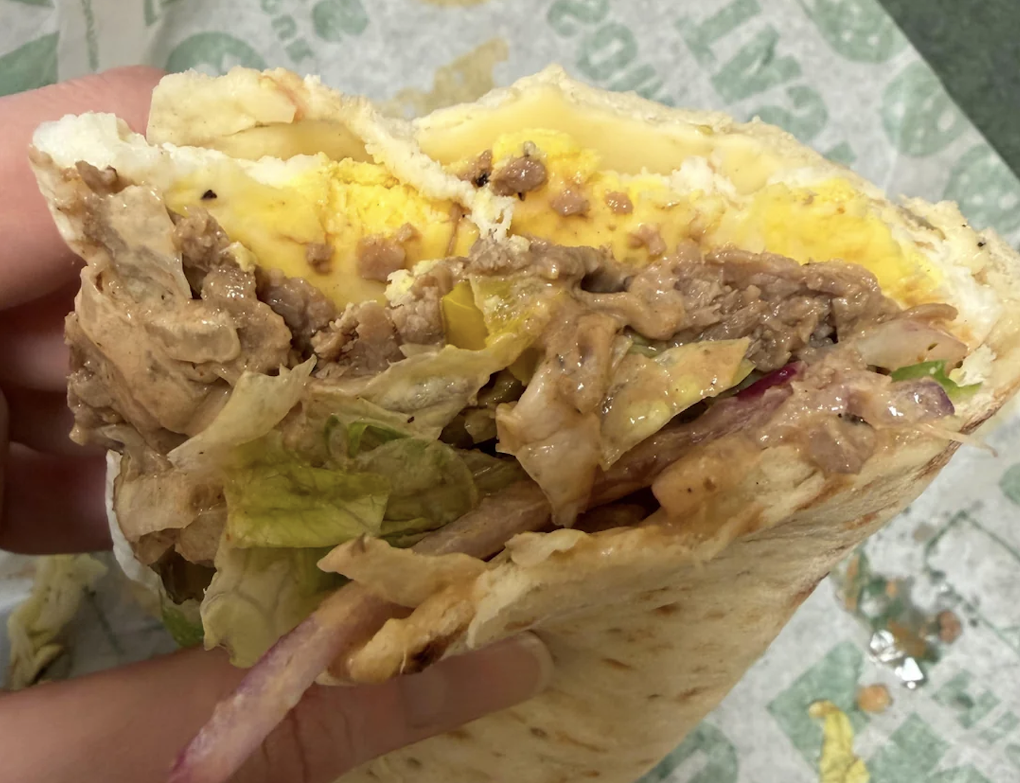 A hand holds a partially eaten wrap filled with beef, lettuce, egg, and sauce. The wrap is on paper with green text in the background.