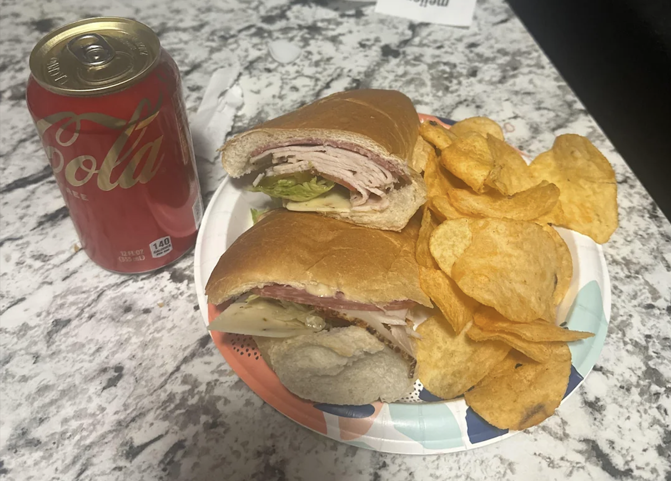 A plate with a sub sandwich, potato chips, and a can of Coca-Cola on a marbled countertop.