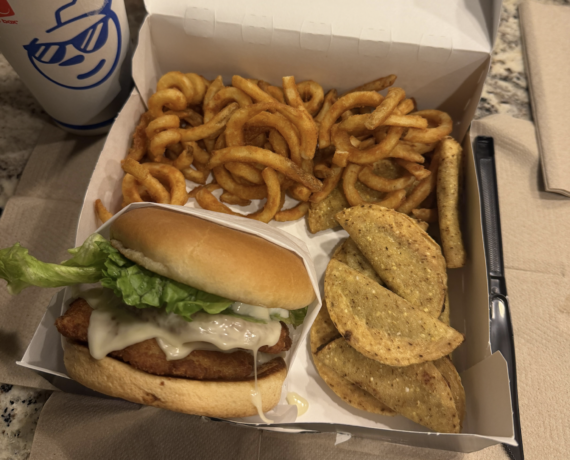 A takeout meal with a chicken sandwich topped with lettuce and cheese, curly fries, potato wedges, a cup with a straw, and a black plastic knife on a napkin, all on a counter.
