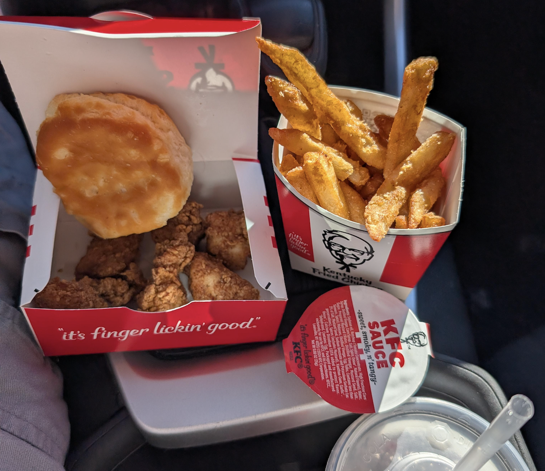 KFC meal with a biscuit, chicken tenders, seasoned fries in a red box, a cup of sauce, and a drink in a plastic cup with a straw on a car armrest.