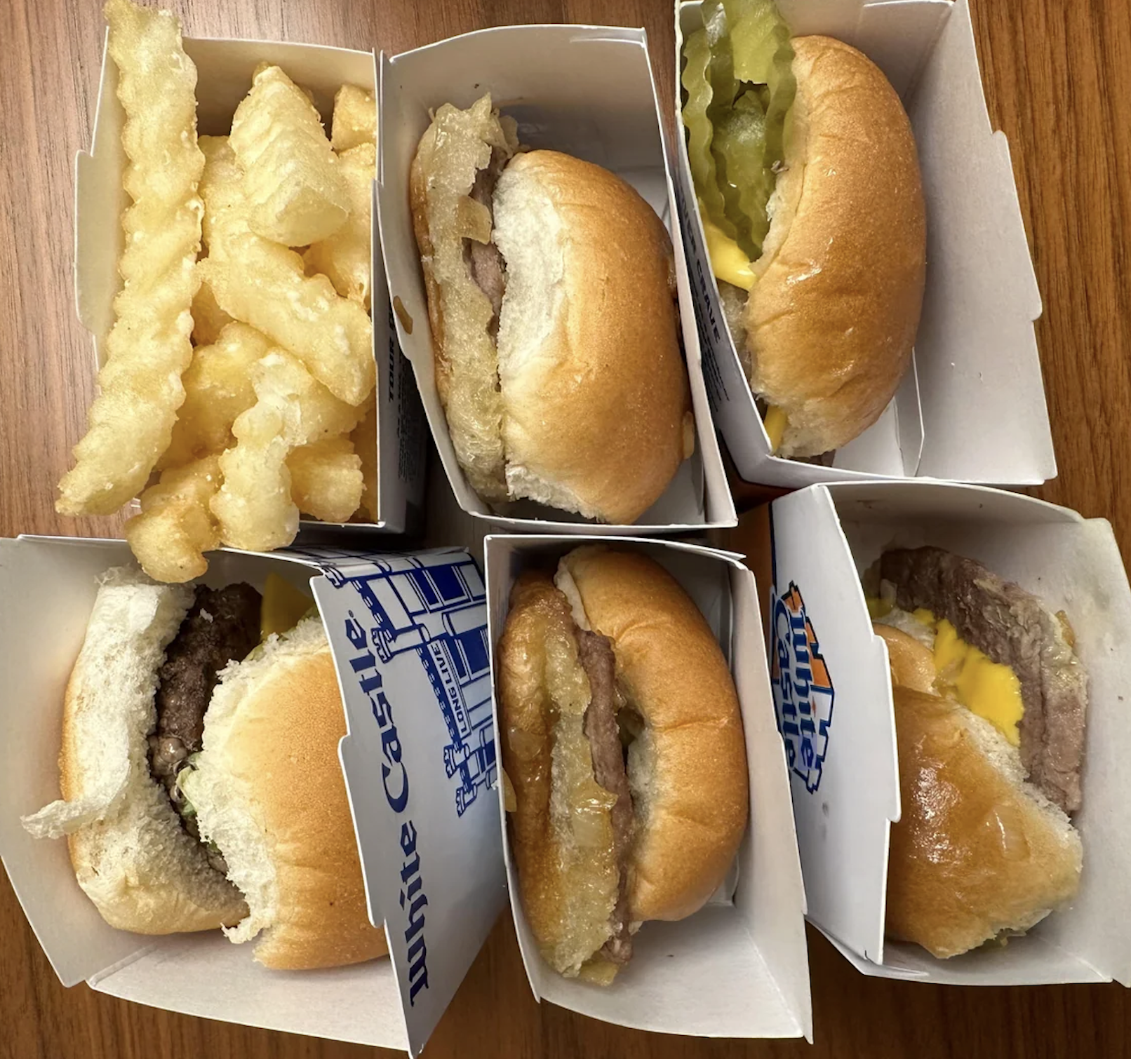 Six White Castle sliders with crinkle-cut fries in a box, arranged on a wooden table. Some sliders have cheese, pickles, or onions visible in their buns.