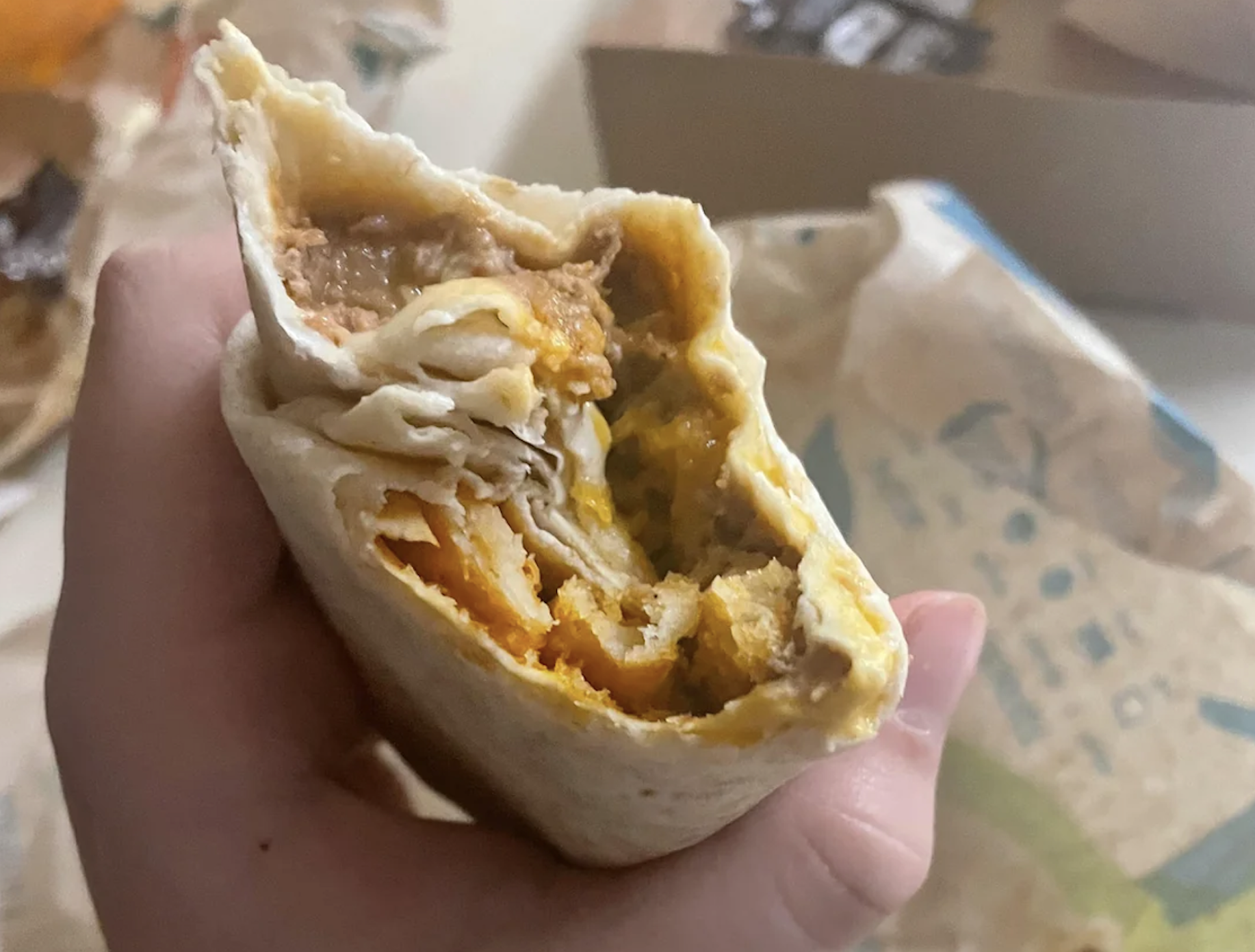 A hand holds a partially eaten burrito filled with various ingredients including cheese, beans, and possibly meat. Wrapping paper and a food container are visible in the background.