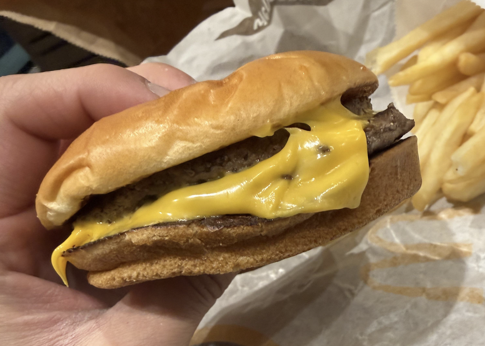 A hand holds a cheeseburger with melted cheese and a beef patty in a soft bun. Crinkle-cut fries are visible in the background on a paper wrapper.