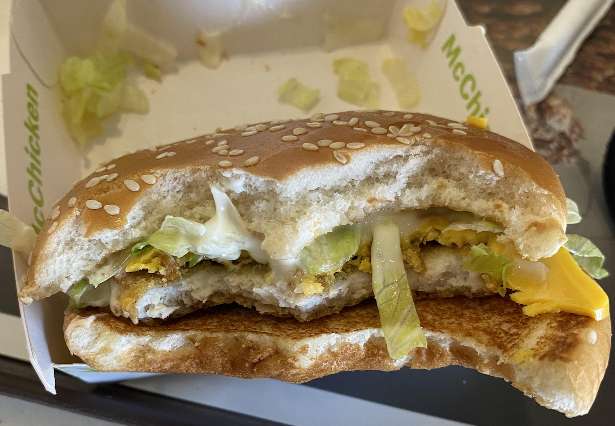 A close-up of a half-eaten chicken sandwich with lettuce, cheese, and mayonnaise in a sesame seed bun, resting in a takeout box.