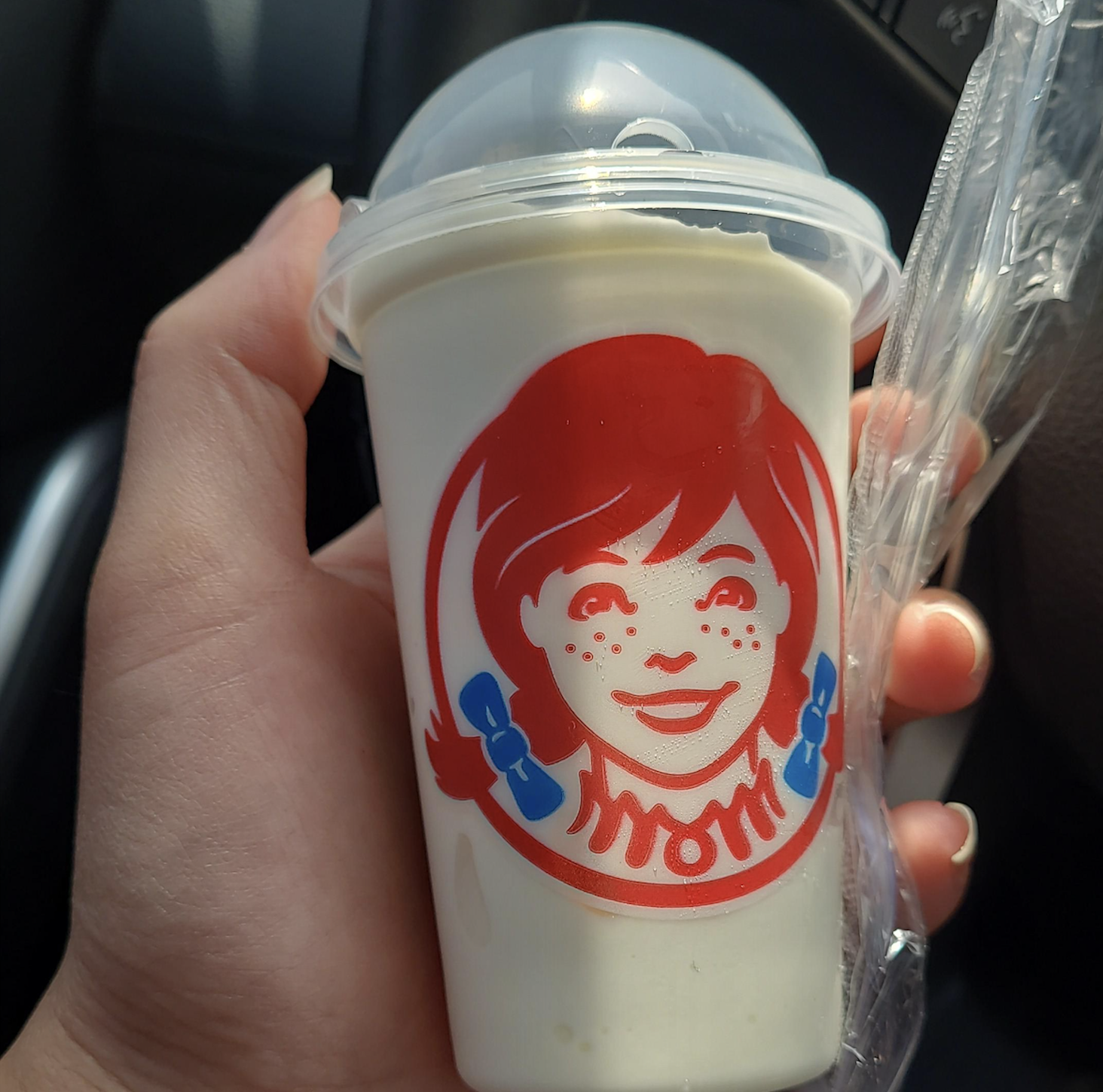 A hand holds a Wendy’s vanilla Frosty in a clear cup with a dome lid, displaying the red logo of a smiling girl with pigtails and blue bows.