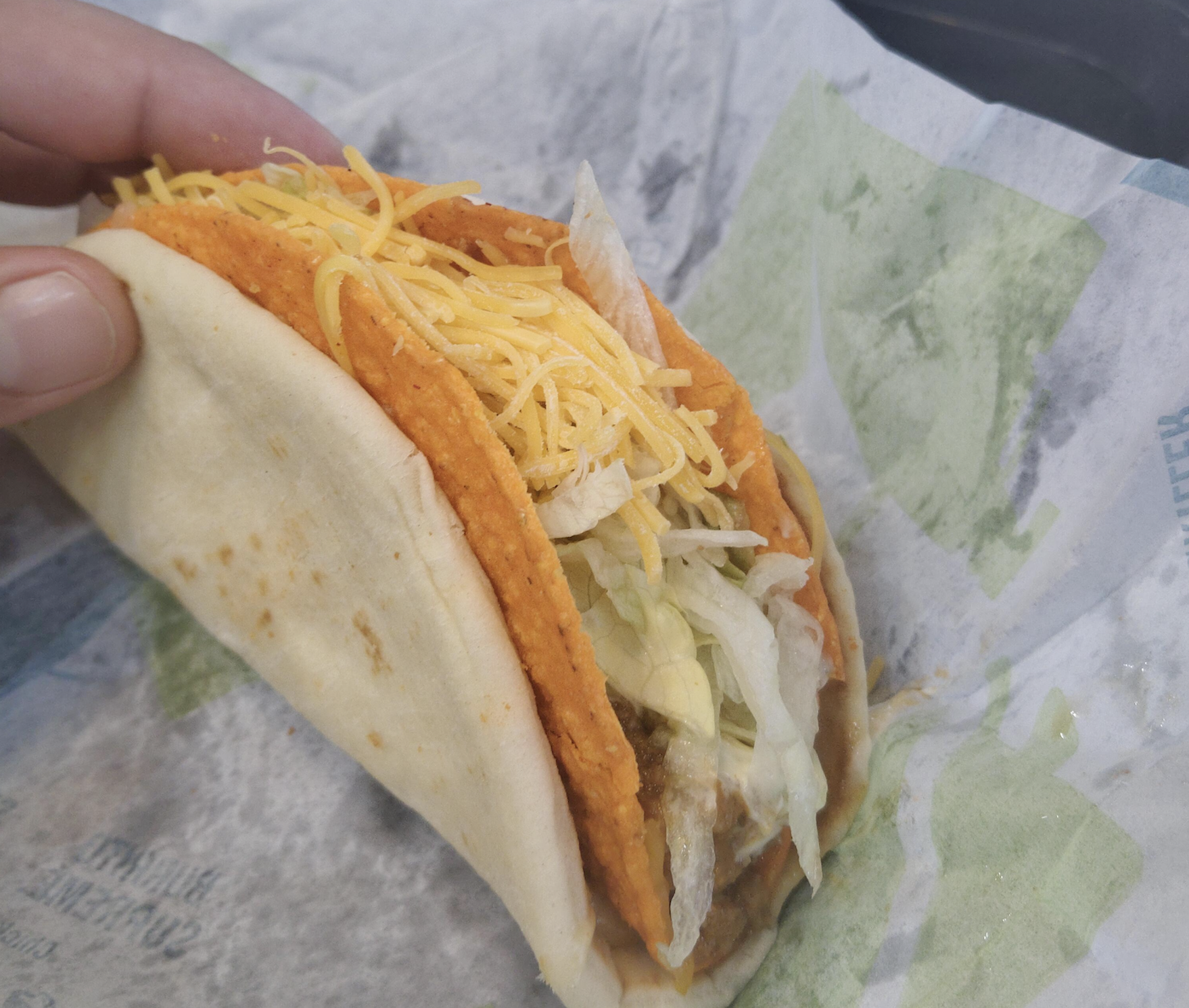 A hand holds a taco with shredded cheddar cheese, lettuce, and a crunchy orange shell inside a soft tortilla, resting on branded fast food paper.
