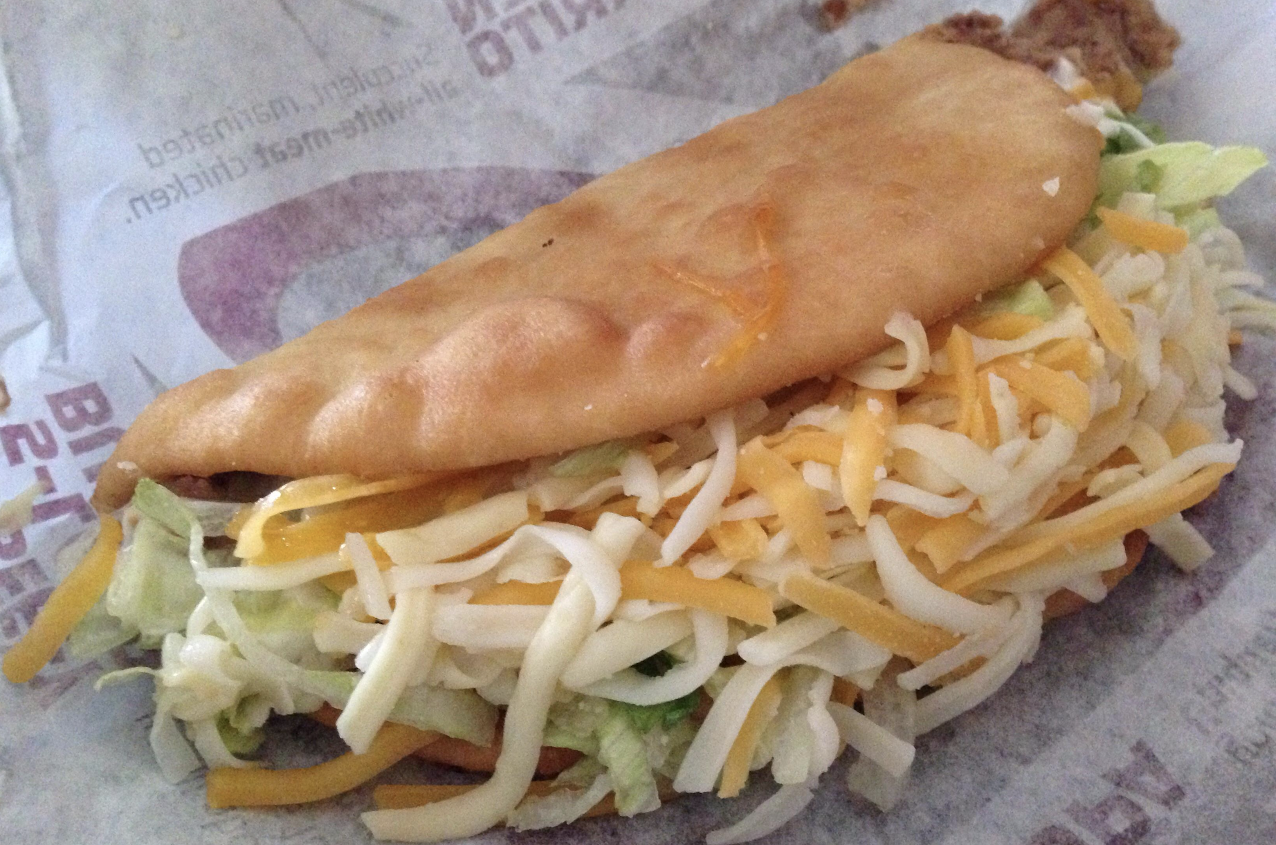 A close-up of a taco with a fried shell, filled with shredded lettuce and a mix of yellow and white shredded cheese, on a fast-food wrapper.