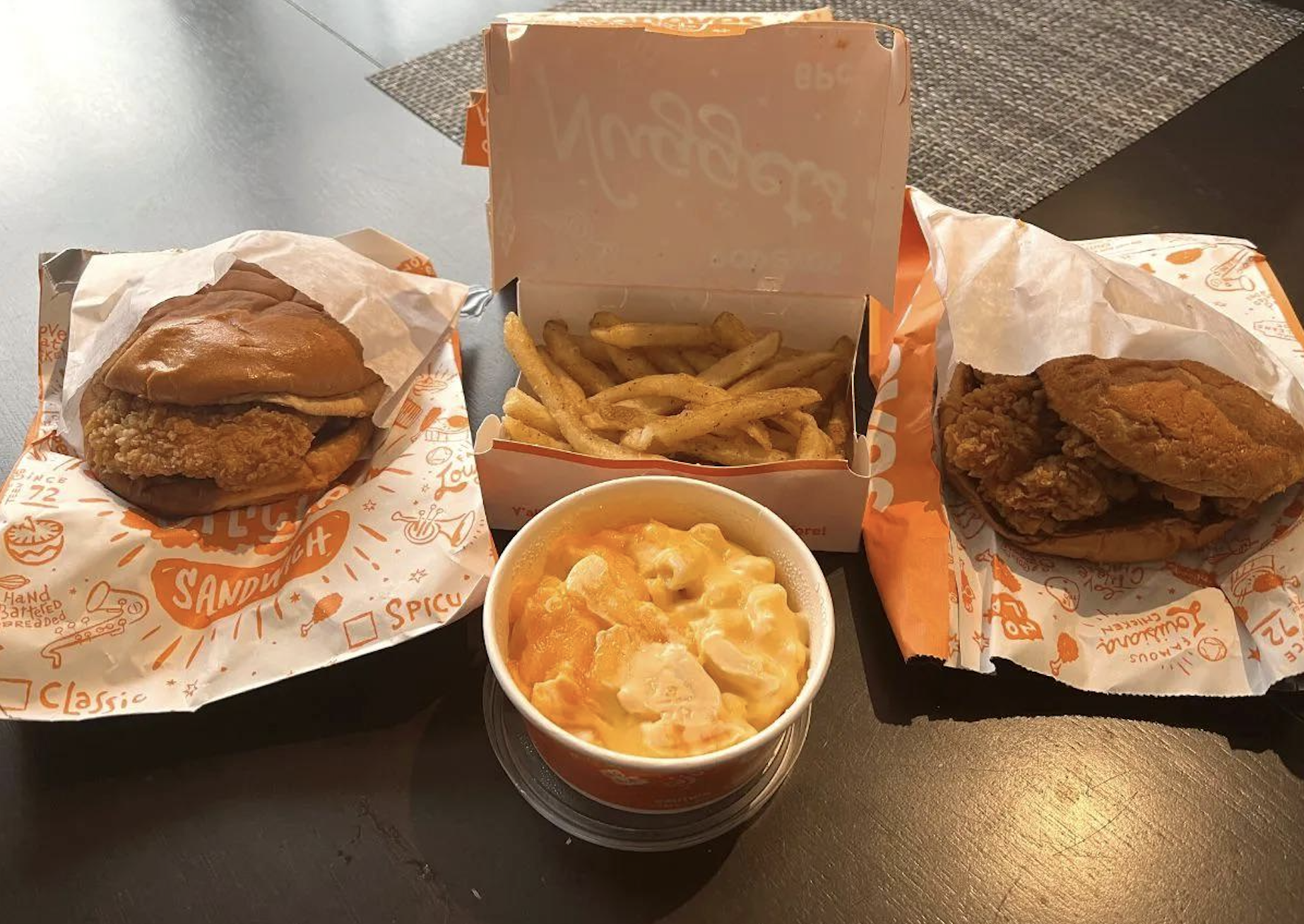 Two fried chicken sandwiches, a cup of macaroni and cheese, and a box of French fries are arranged on a black table. The food is served in branded orange and white packaging.