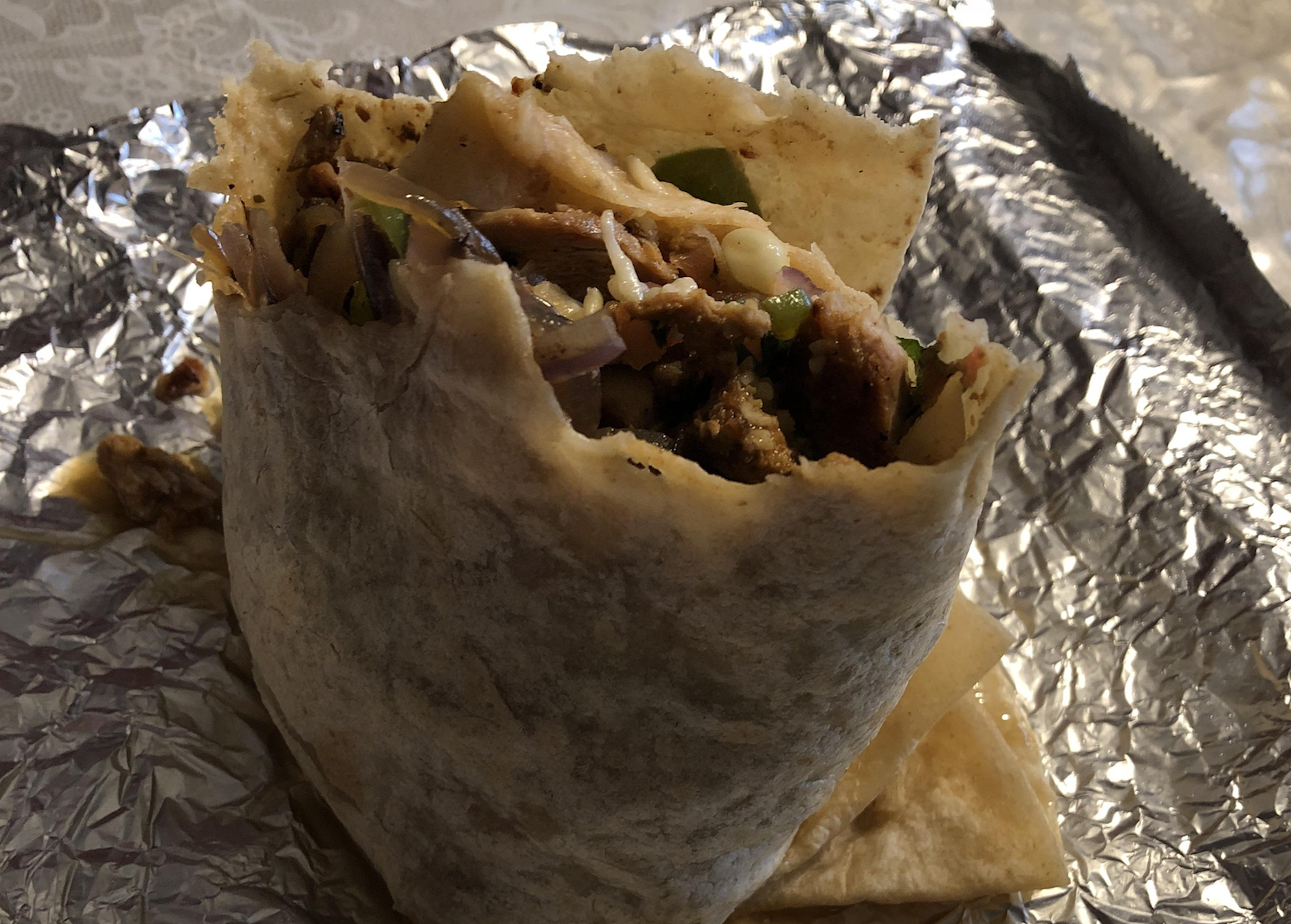 A close-up of a partially unwrapped shawarma wrap with grilled meat, onions, and vegetables, resting on crinkled aluminum foil.