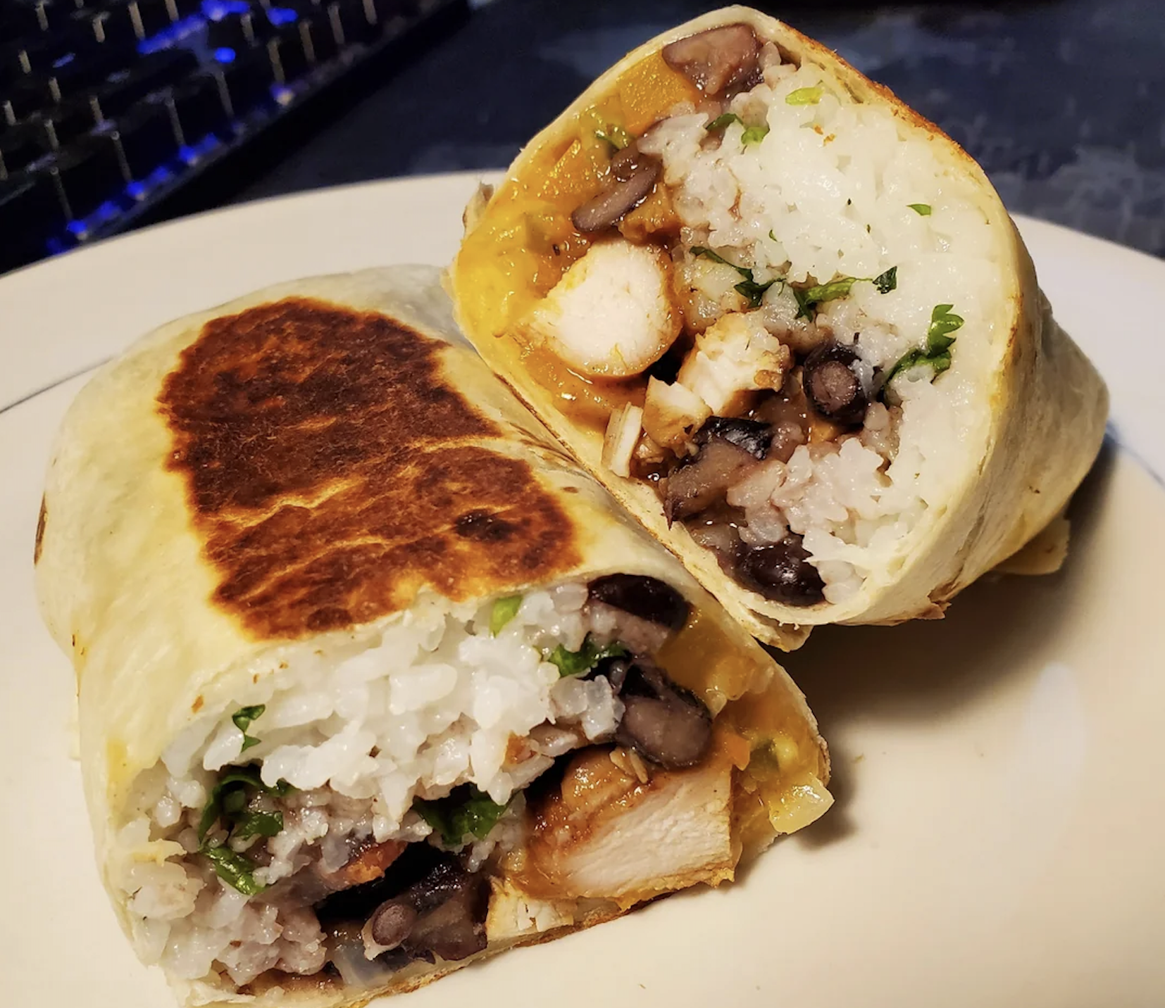 A close-up of a burrito cut in half, showing rice, black beans, chicken, and herbs inside a toasted tortilla on a white plate.