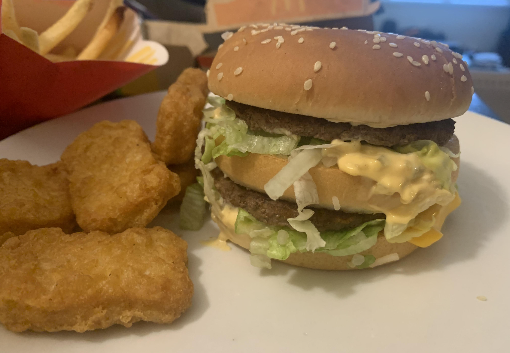 A sesame seed burger bun with lettuce, cheese, sauce, and two beef patties sits on a plate next to three breaded chicken nuggets. French fries are in the background.