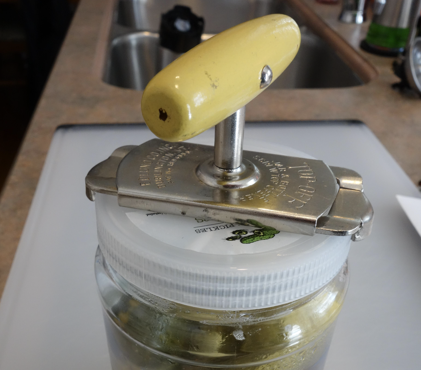 A jar opener with a yellow handle is placed on top of a jar with a white lid, set on a kitchen counter near a sink.