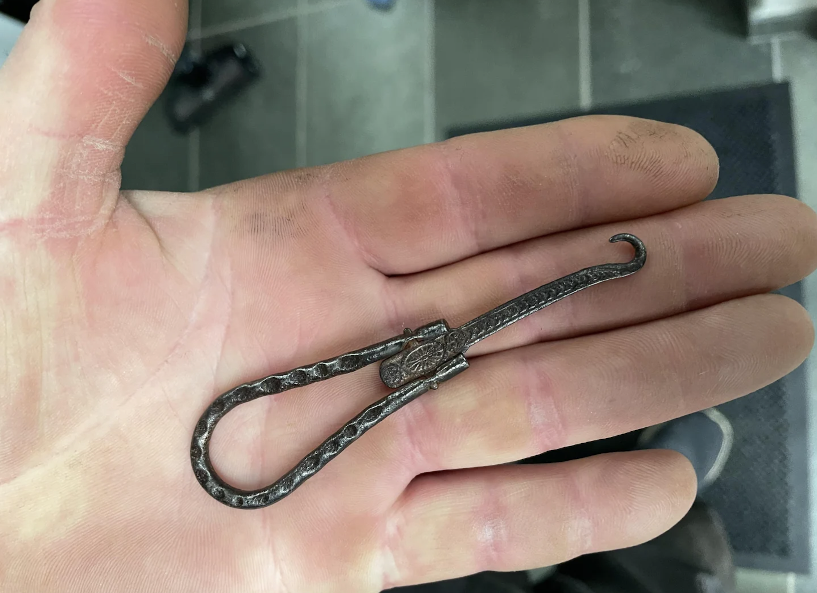 A person’s hand holds a small, twisted metal tool with a hooked end, resembling a miniature ironwork piece. The hand appears slightly dirty, possibly from manual work.
