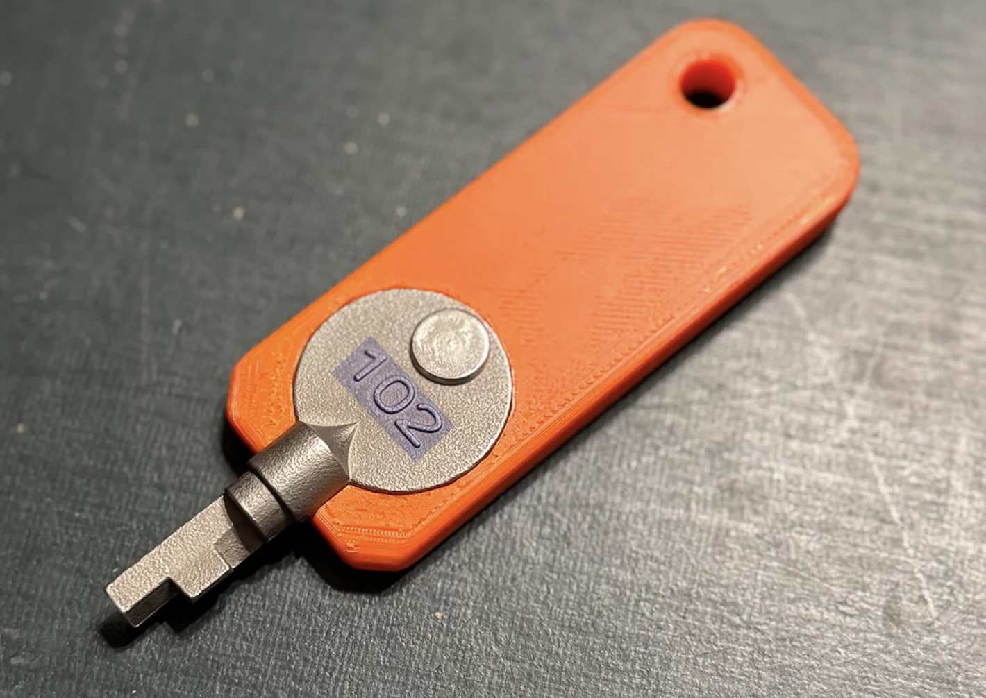 A metallic key with a flat, unusual tip is attached to an orange plastic tag labeled “102.” The tag has a hole on one end, possibly for attaching to a keyring.