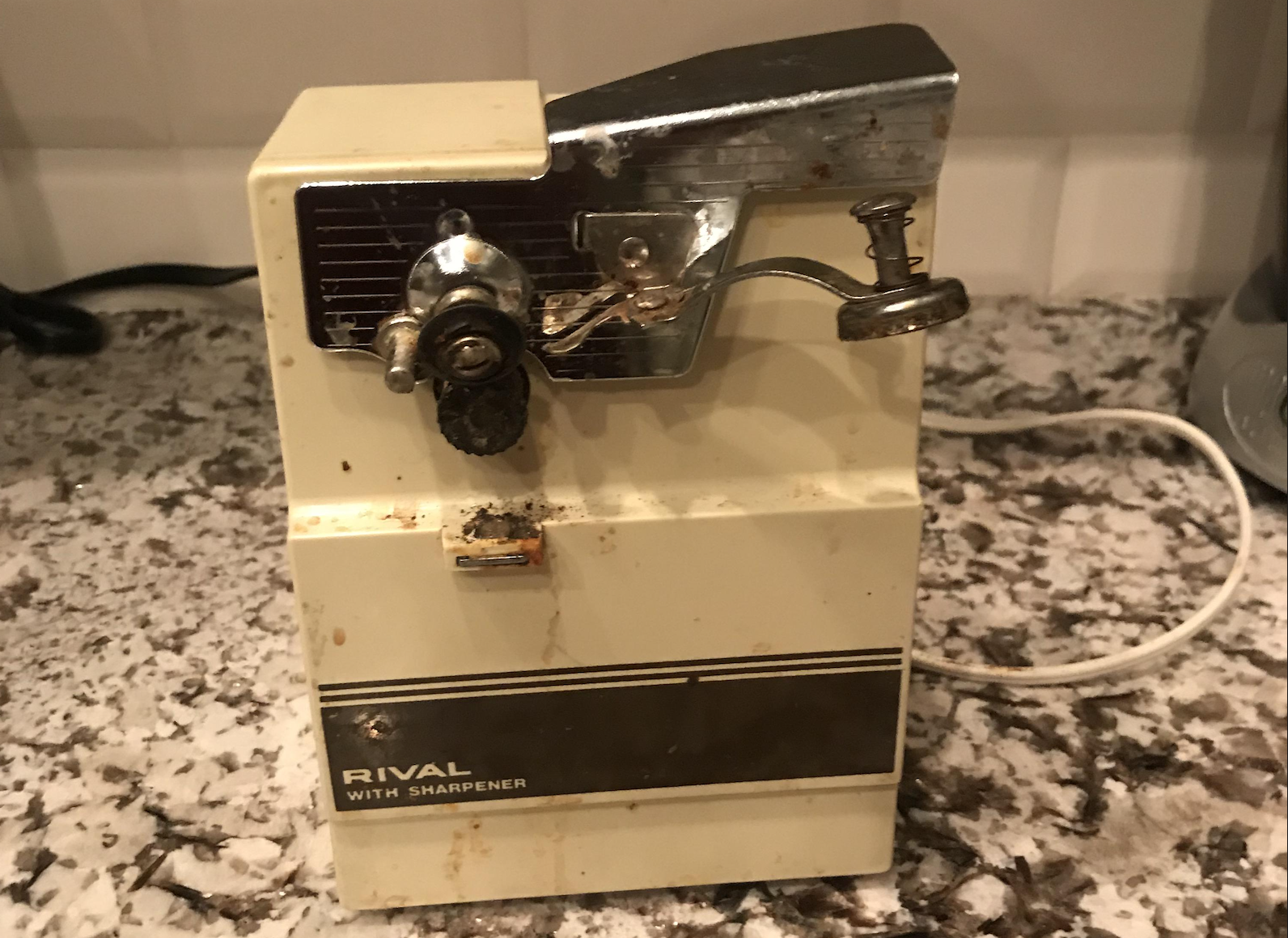 A vintage electric can opener with a built-in sharpener, labeled “Rival With Sharpener,” sits on a speckled countertop. The device shows signs of rust and wear.