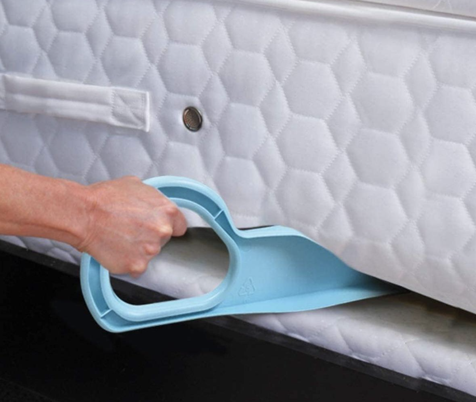 A hand uses a blue plastic tool to lift the corner of a white mattress, revealing the underlying bed frame.