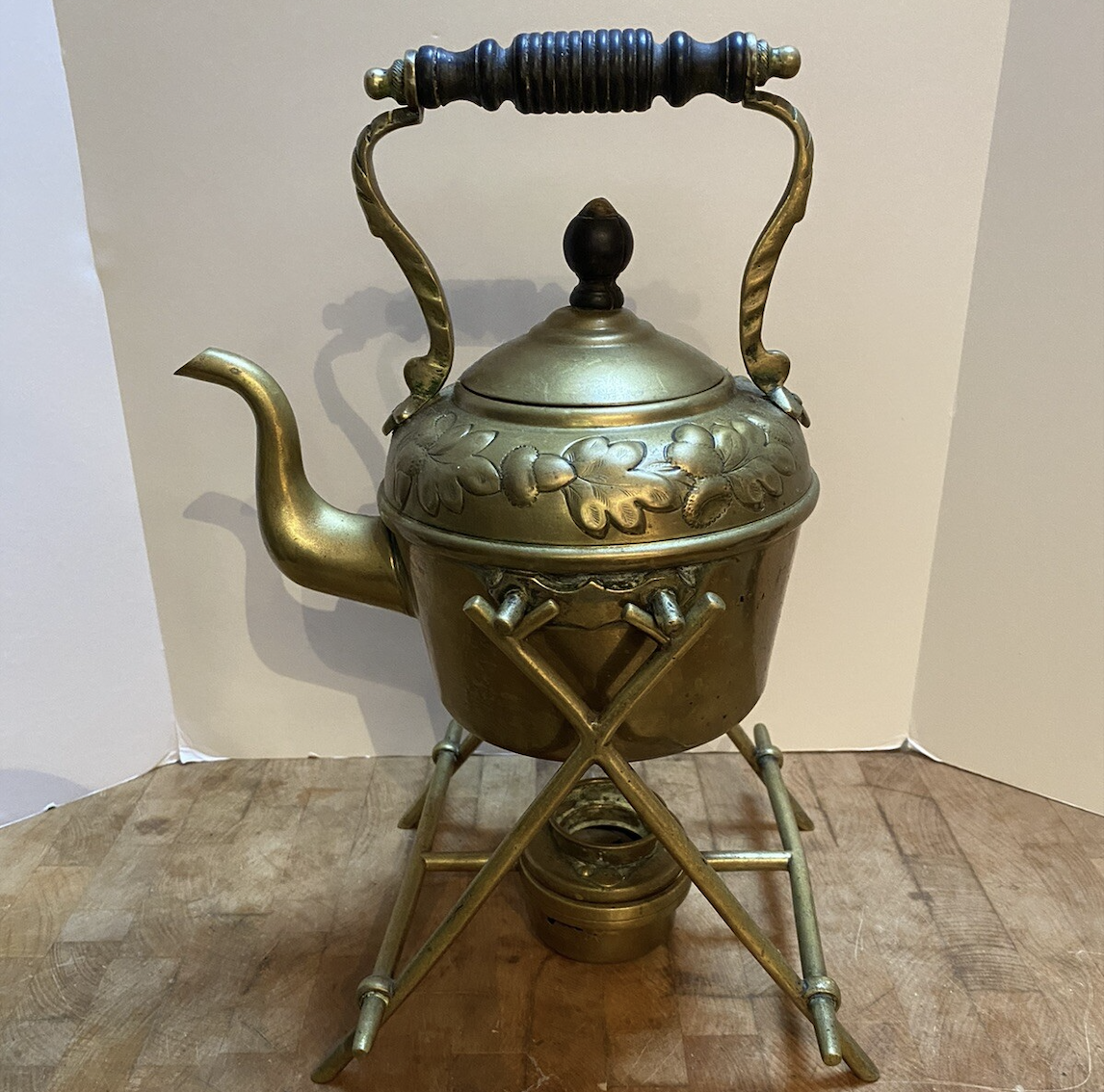Brass teapot with ornate handle and lid, floral design, resting on a matching stand with a small burner beneath, all placed on a wooden surface against a light background.