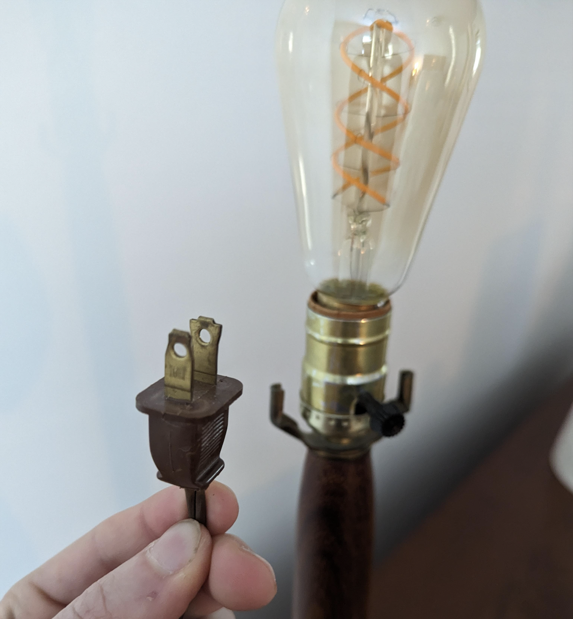 A hand holds a detached electrical plug near a vintage-style light bulb and lamp base, suggesting the lamp needs repair or rewiring.