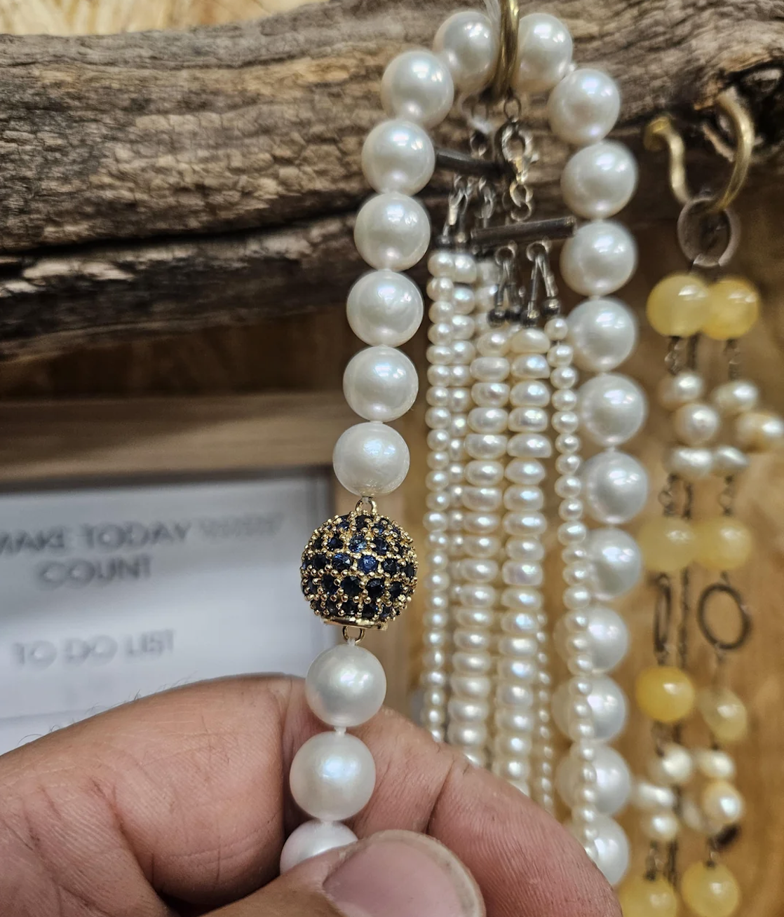 A hand holds a pearl necklace with a decorative clasp featuring dark blue and gold accents. Other pearl necklaces hang in the background, with a wooden piece and a blurred sign visible.