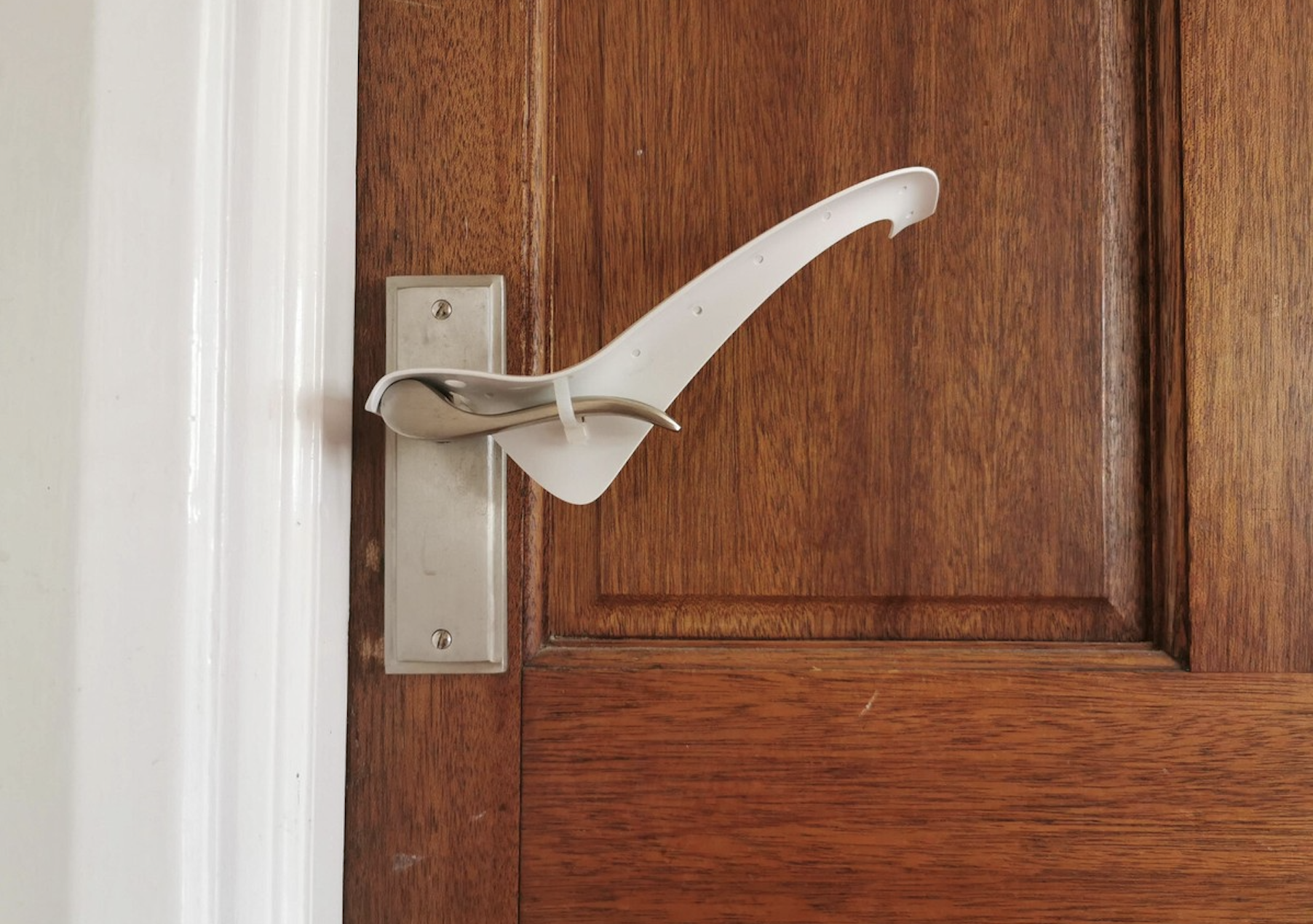 A plastic device is attached to a door handle, allowing the door to be opened with an arm instead of a hand. The wooden door has a silver plate, and the device is mounted horizontally.