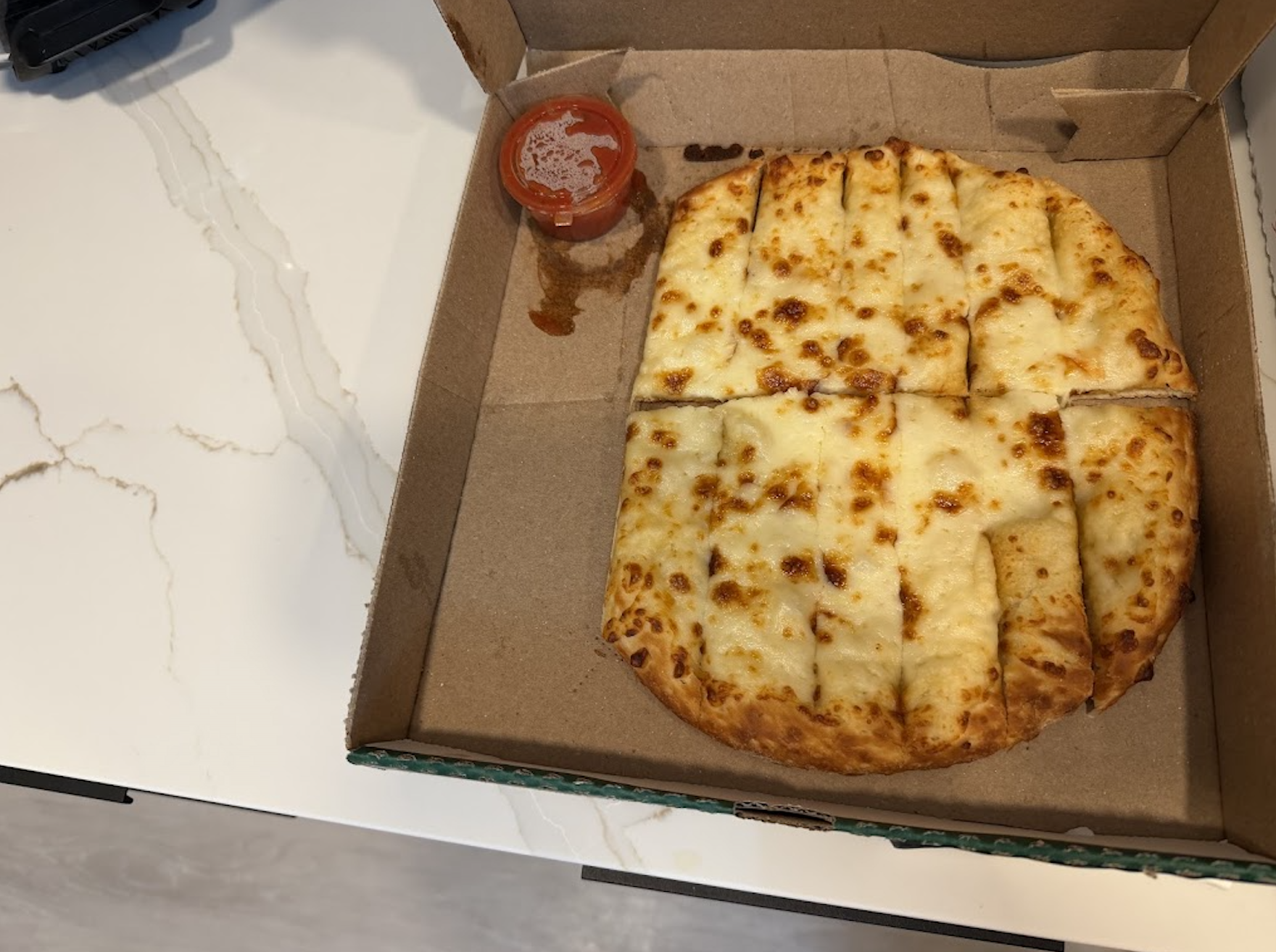 An open pizza box with cheesy breadsticks cut into squares and a small container of marinara sauce on a white countertop.