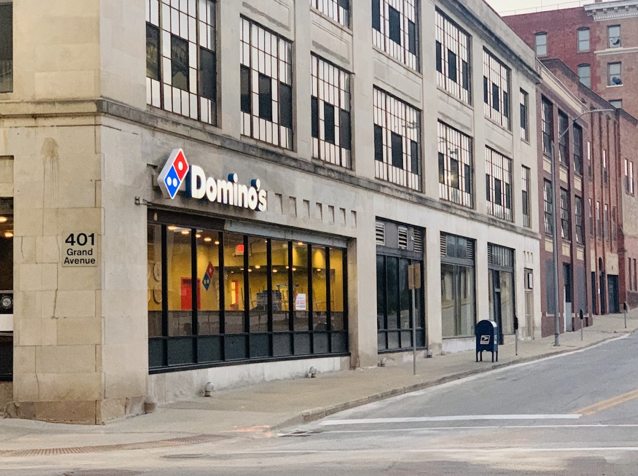 A Domino’s pizza restaurant is on the ground floor of a large, beige building at 401 Grand Avenue. The street is quiet with a mailbox nearby and few signs of people or traffic.
