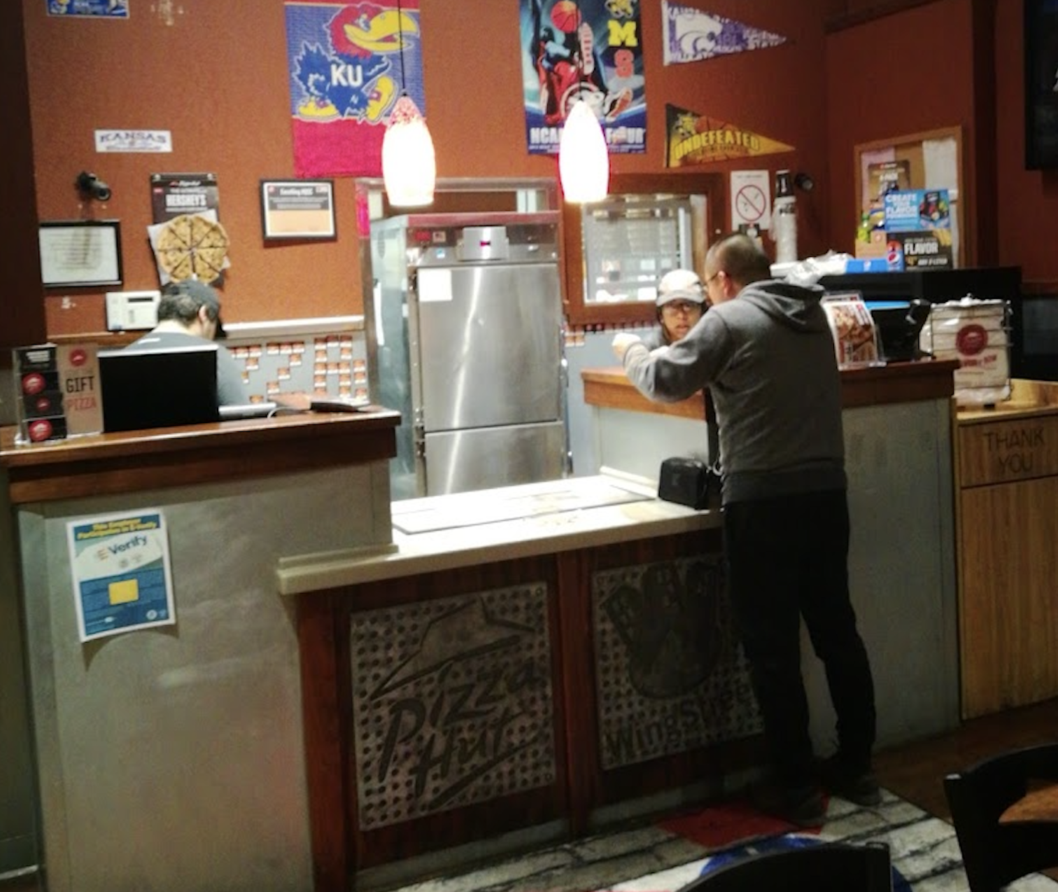 A man stands at a counter inside a Pizza Hut, speaking to an employee. Another worker is at the register. Various posters and signs decorate the back wall. The area is warmly lit with hanging lights.