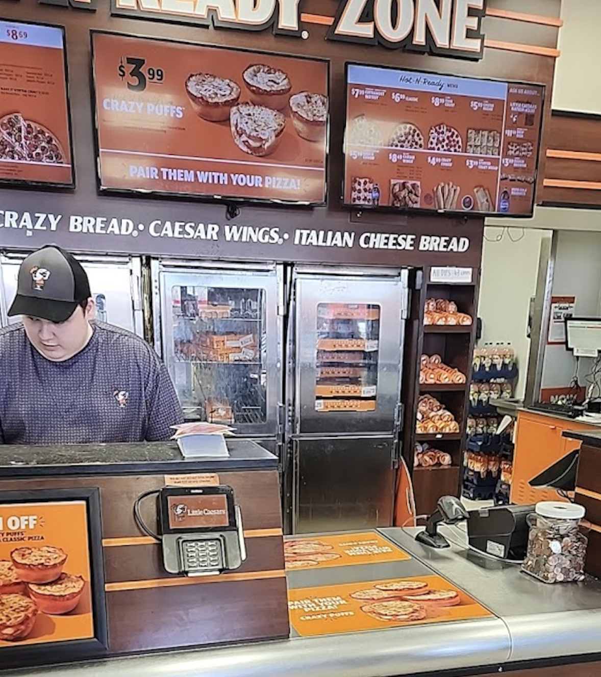 A worker in a pizza shop stands behind the counter. Digital menu screens and pizza ovens are visible in the background, with bread and soda bottles displayed on shelves nearby.