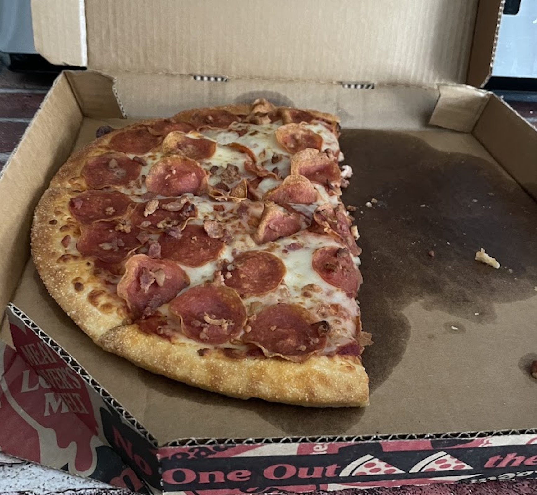 Half of a pepperoni pizza with a thick crust sits in an open cardboard box on a table, with several slices missing and some grease stains visible in the box.