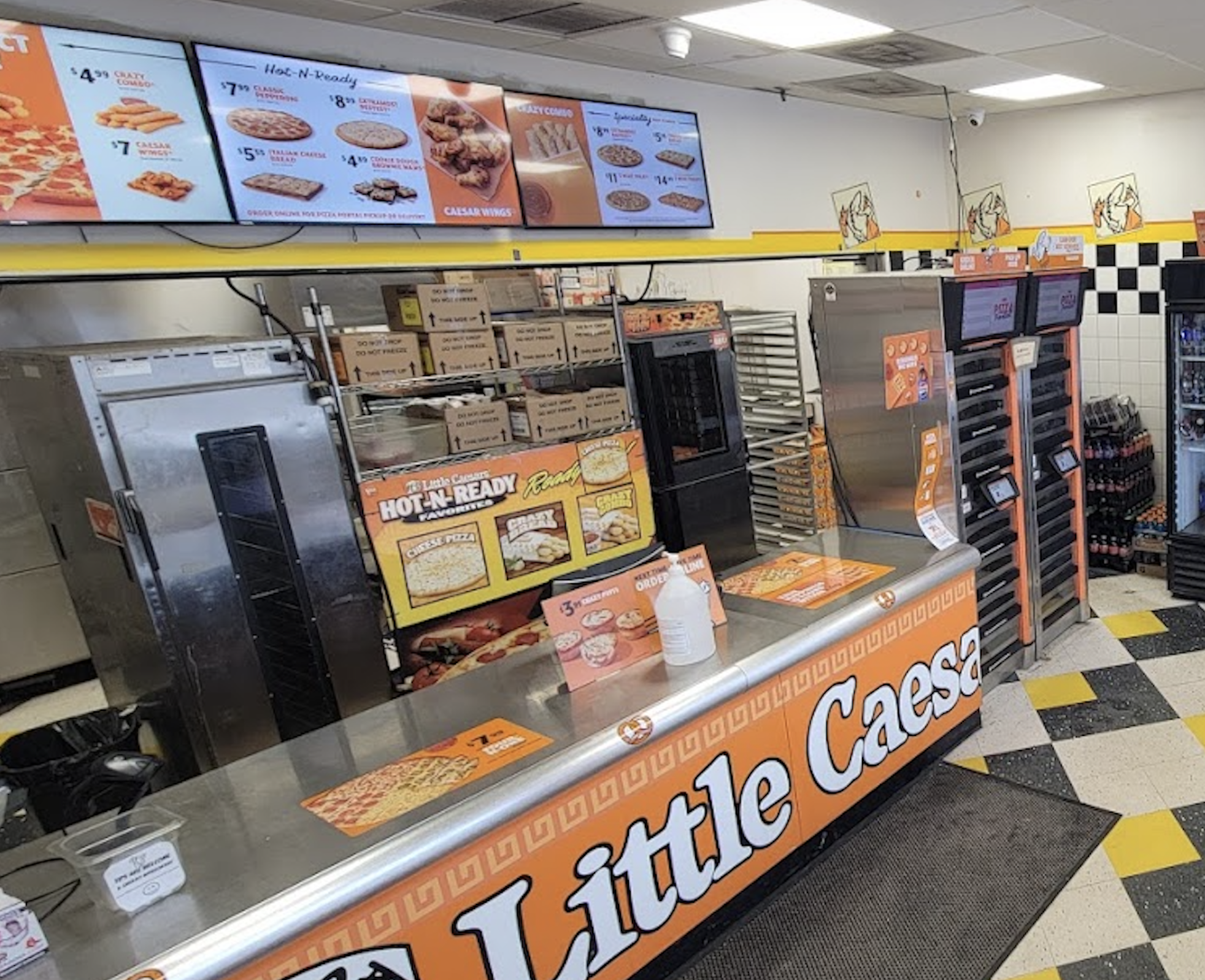 Little Caesars counter with pizza boxes stacked behind, menus on digital screens above, and orange branding visible throughout the store. Sanitizer bottle and promo boards are on the counter.