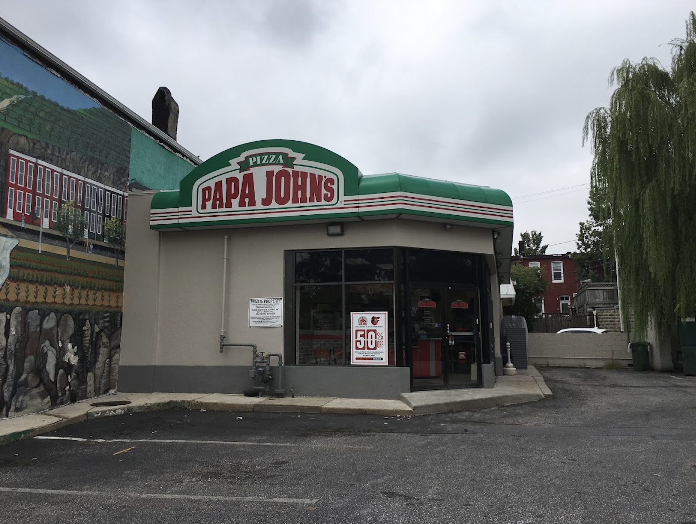 Small Papa John’s pizza restaurant with a green and red sign, located next to a mural and a willow tree. The building is on a mostly empty parking lot under a cloudy sky.