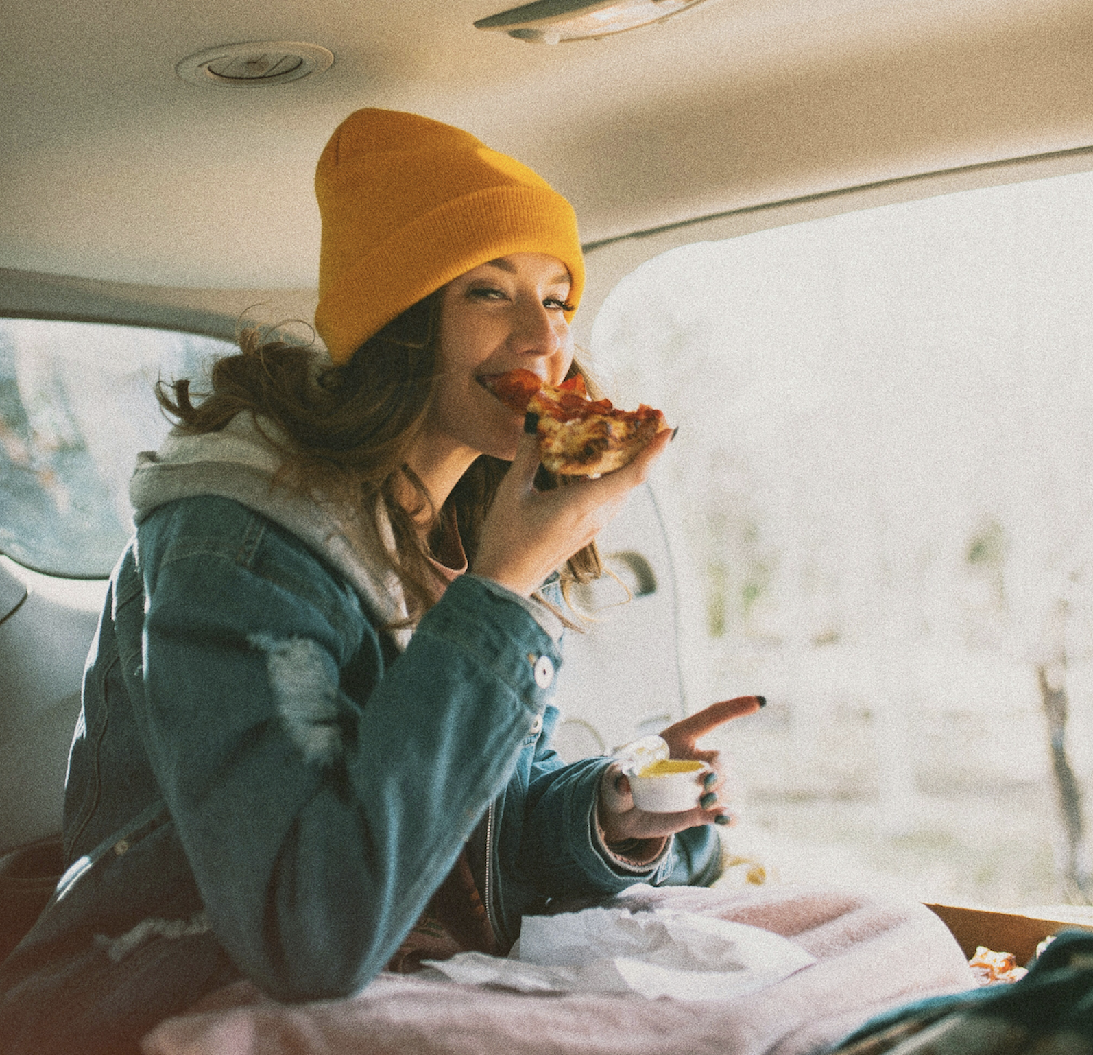 A person wearing a yellow beanie and denim jacket is smiling and eating pizza while sitting in the back of a car with sunlight streaming through the window.