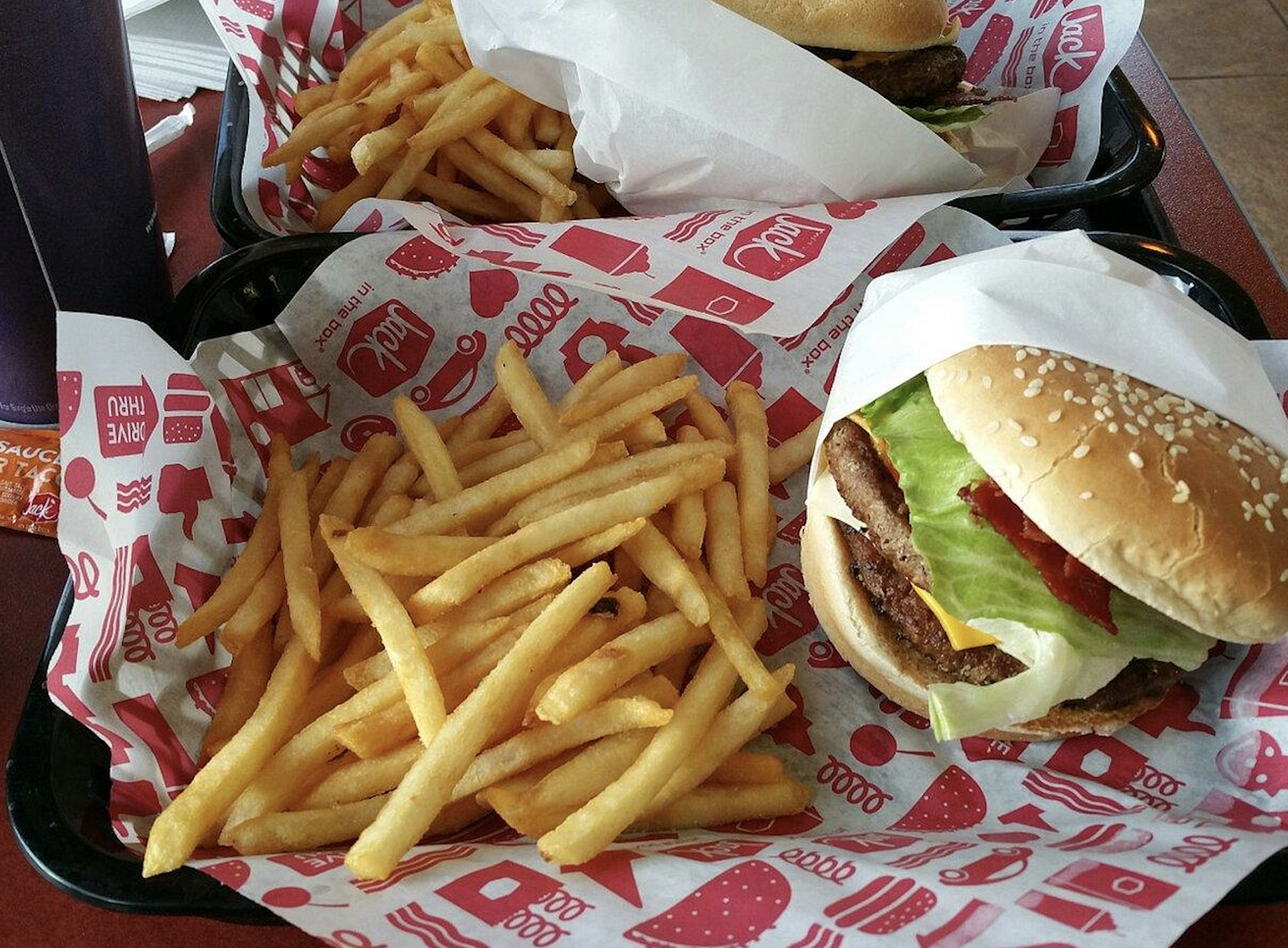 A tray with a burger topped with lettuce and ketchup in a sesame seed bun, served with a large portion of golden French fries on red and white Jack in the Box branded paper. Another similar tray is in the background.