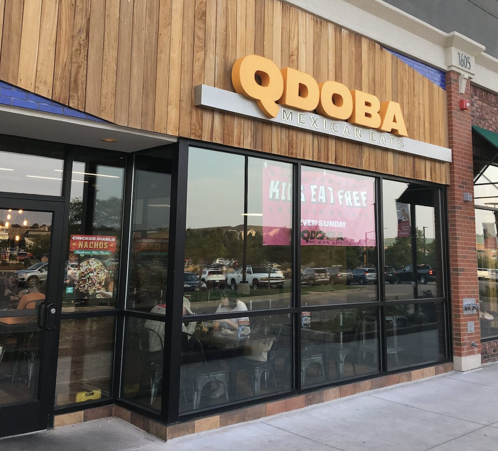 Exterior of a QDOBA Mexican Eats restaurant with large windows, a wooden facade, and a pink sign in the window that says “KIDS EAT FREE EVERY SUNDAY.” Some people are visible sitting inside.