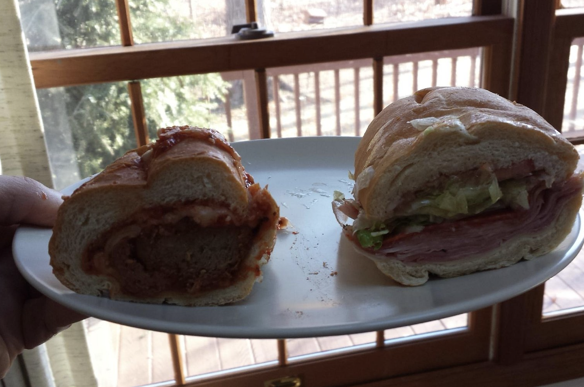 A white plate holds two sandwich halves: one is a meatball sub with sauce, the other has deli meats, cheese, and lettuce. A window and wooden deck are visible in the background.