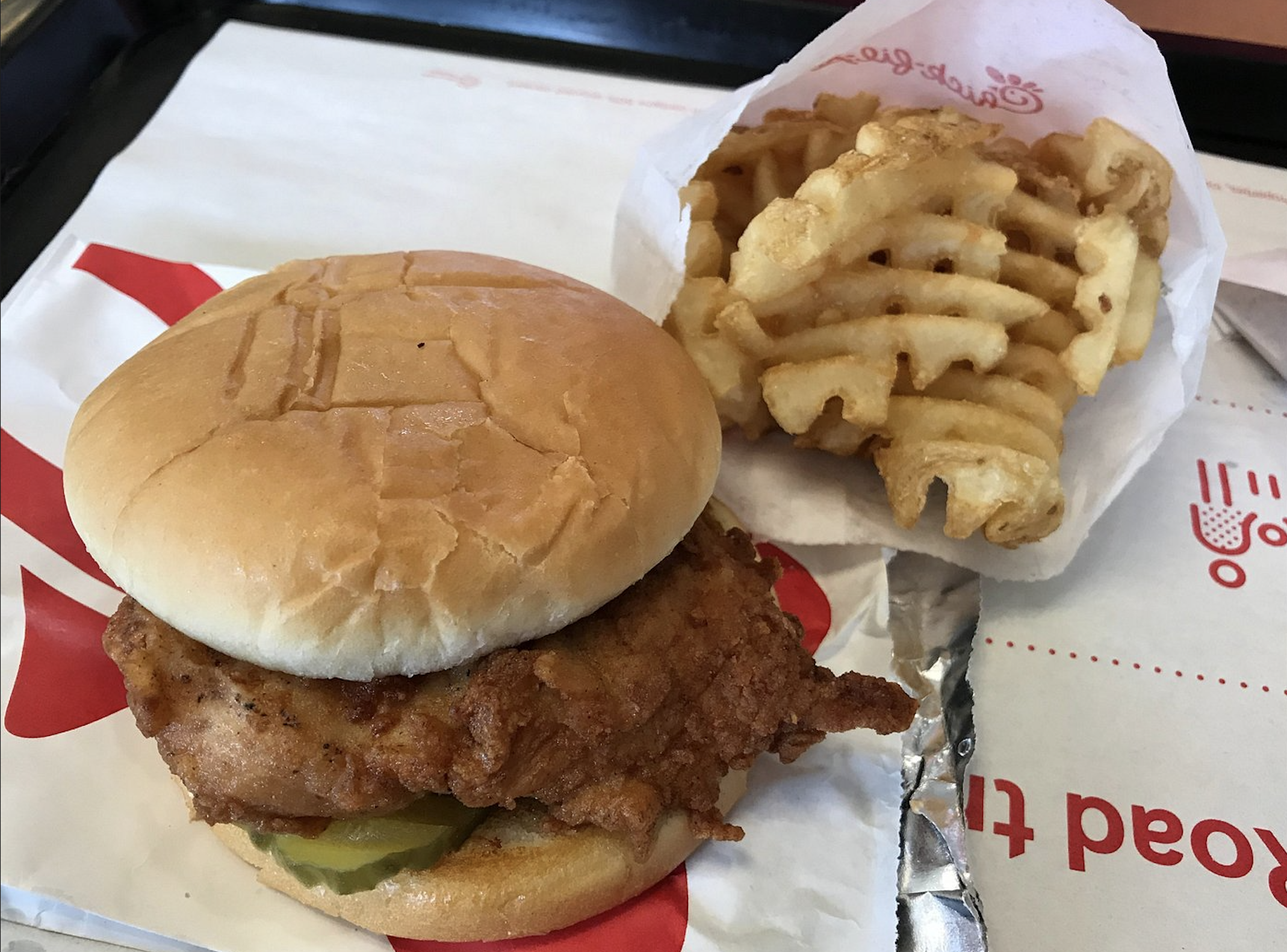 A fried chicken sandwich with pickles on a bun sits next to a small serving of waffle fries in a paper pouch on a fast food tray.