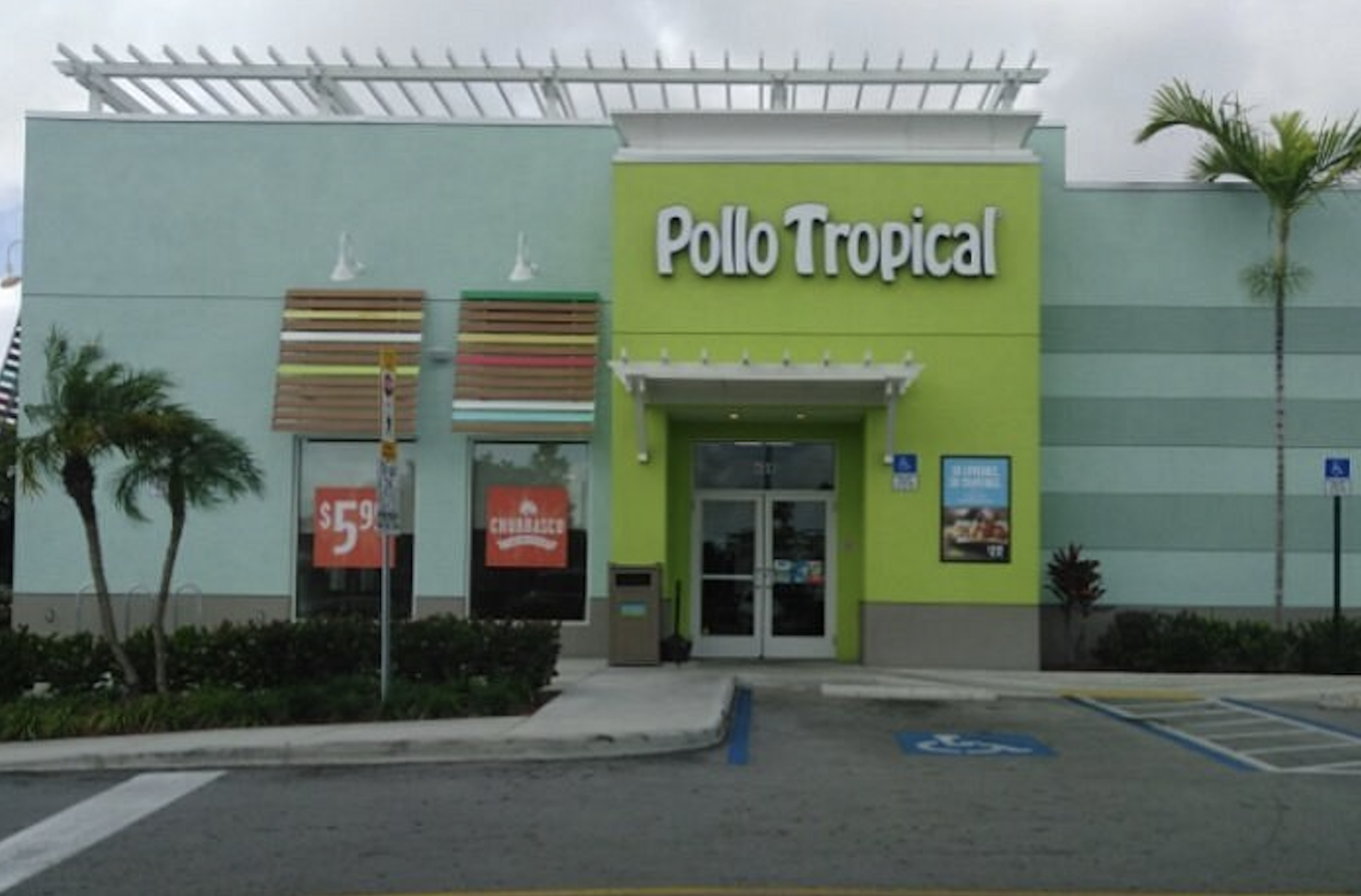 Pollo Tropical restaurant exterior with green and blue walls, large sign, palm tree, parking lot, and posters advertising meal deals in the front windows.