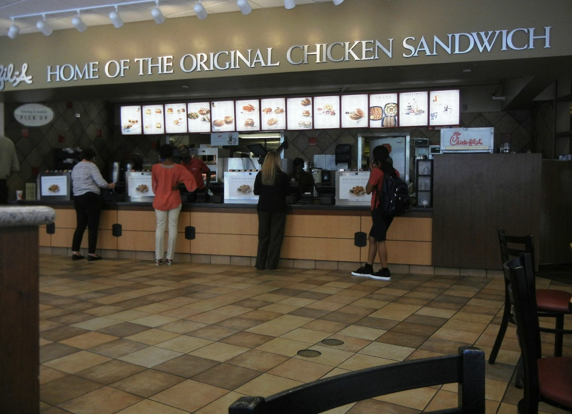 Customers stand in line at a fast food restaurant counter under a sign reading “Home of the Original Chicken Sandwich,” waiting to place their orders.