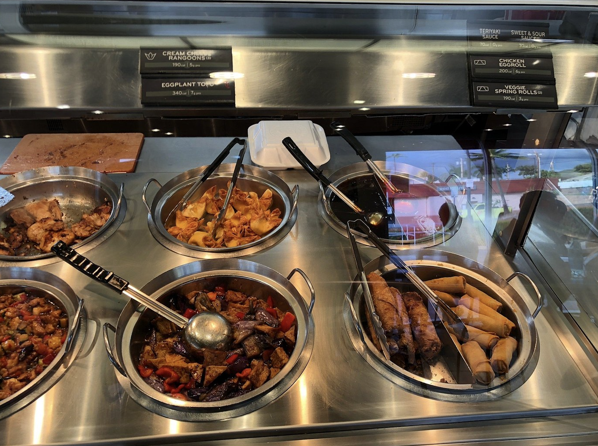 A buffet with six metal trays filled with various Asian dishes, including stir-fries, egg rolls, and spring rolls. Serving utensils are in each tray, and a takeout box sits behind the food.
