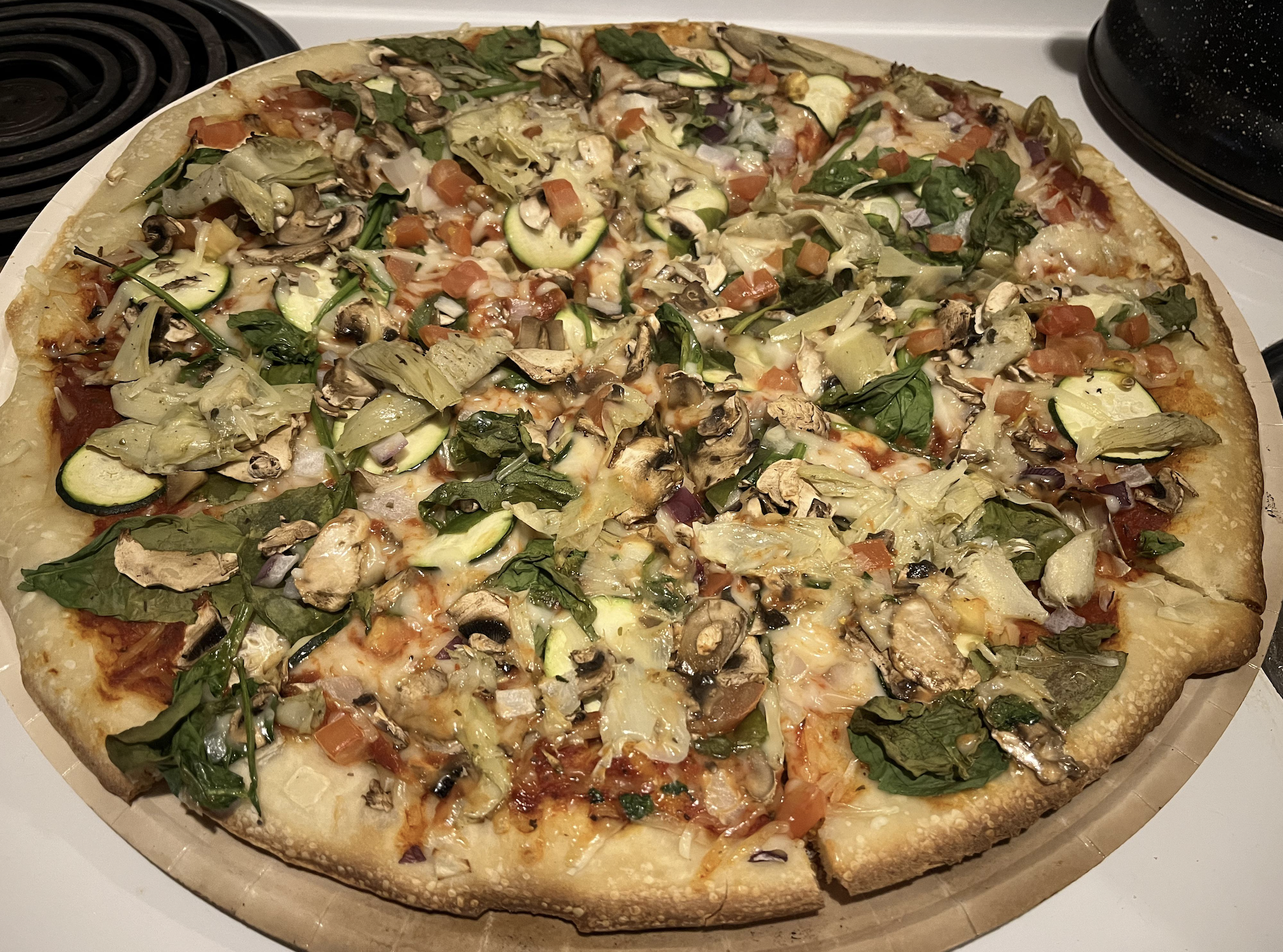 A homemade vegetable pizza topped with spinach, mushrooms, zucchini, tomatoes, artichoke hearts, onions, and melted cheese sits on a round pizza stone beside a stovetop burner.