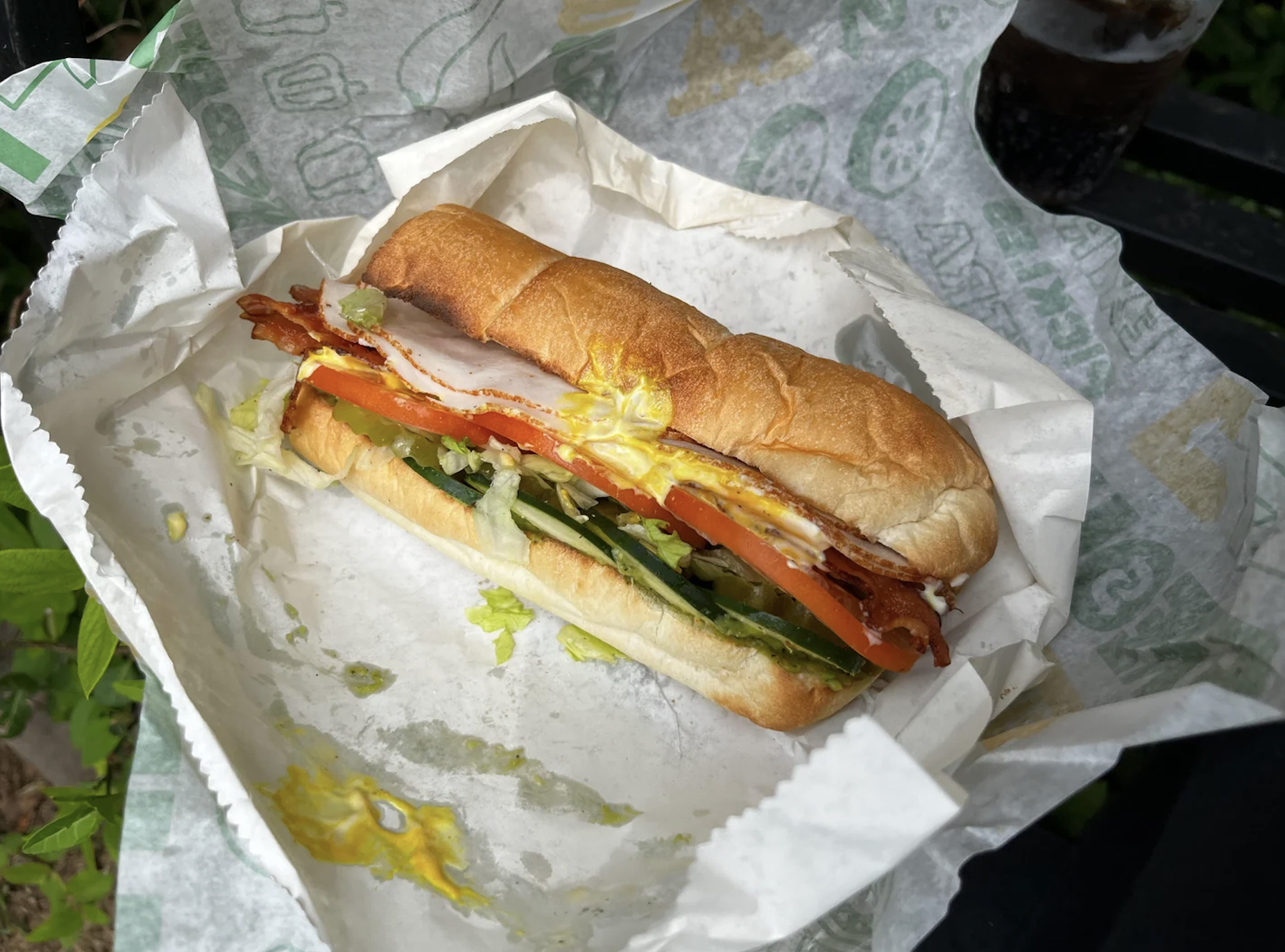 A sub sandwich with turkey, bacon, lettuce, tomato, pickles, and mustard sits on sandwich shop paper. Part of a drink in a cup is visible in the corner.