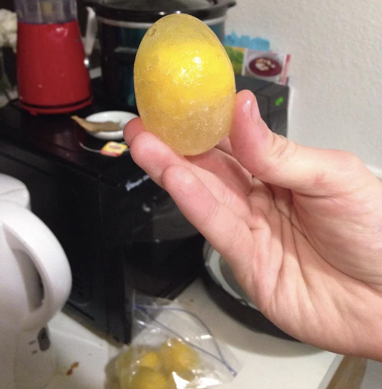 A hand holds a frozen lemon encased in clear ice, with kitchen appliances and a plastic bag on the counter in the background.