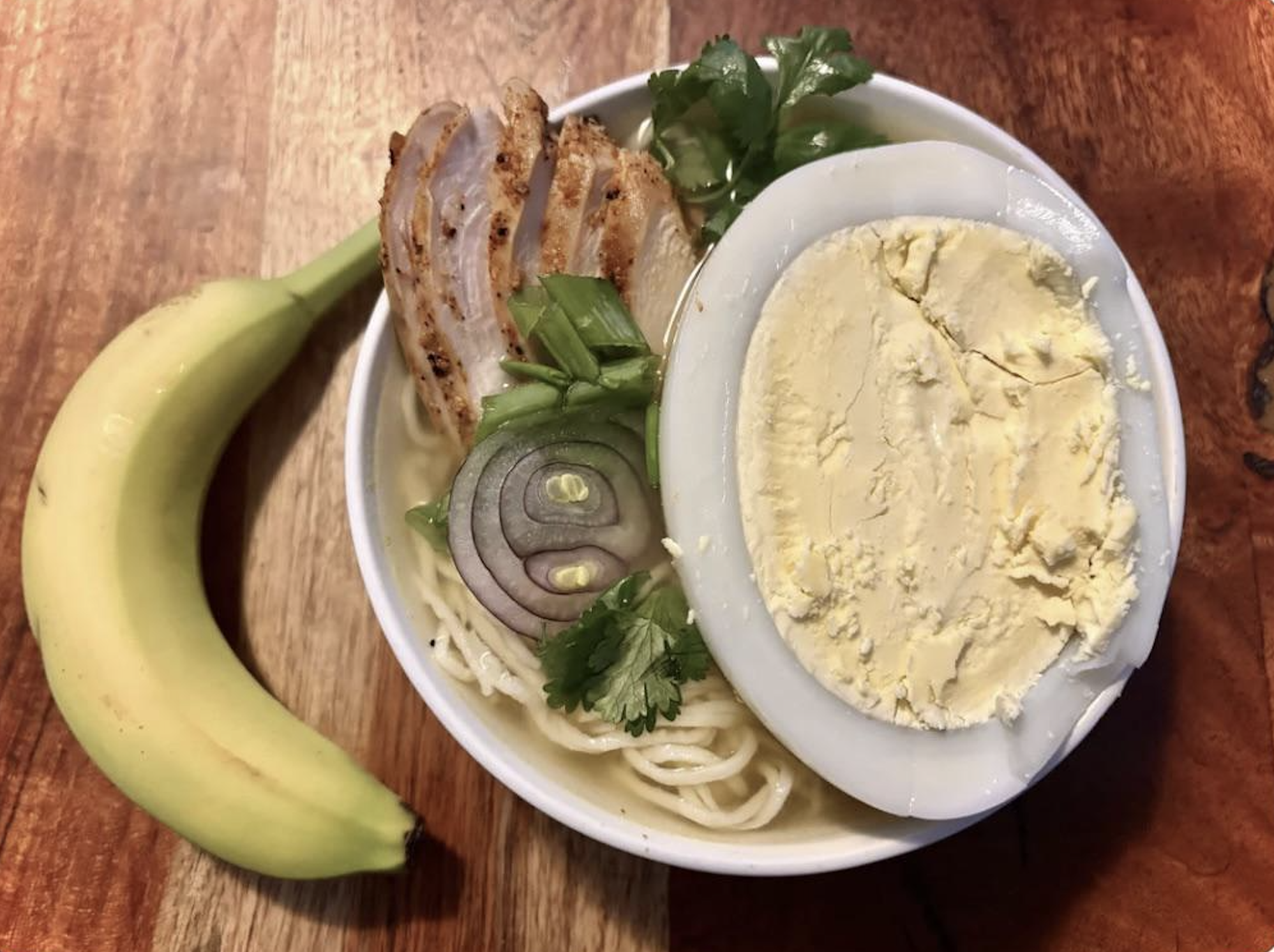 A bowl of noodle soup topped with a halved boiled egg, grilled chicken, sliced red onion, and herbs, sits next to a ripe banana on a wooden surface.