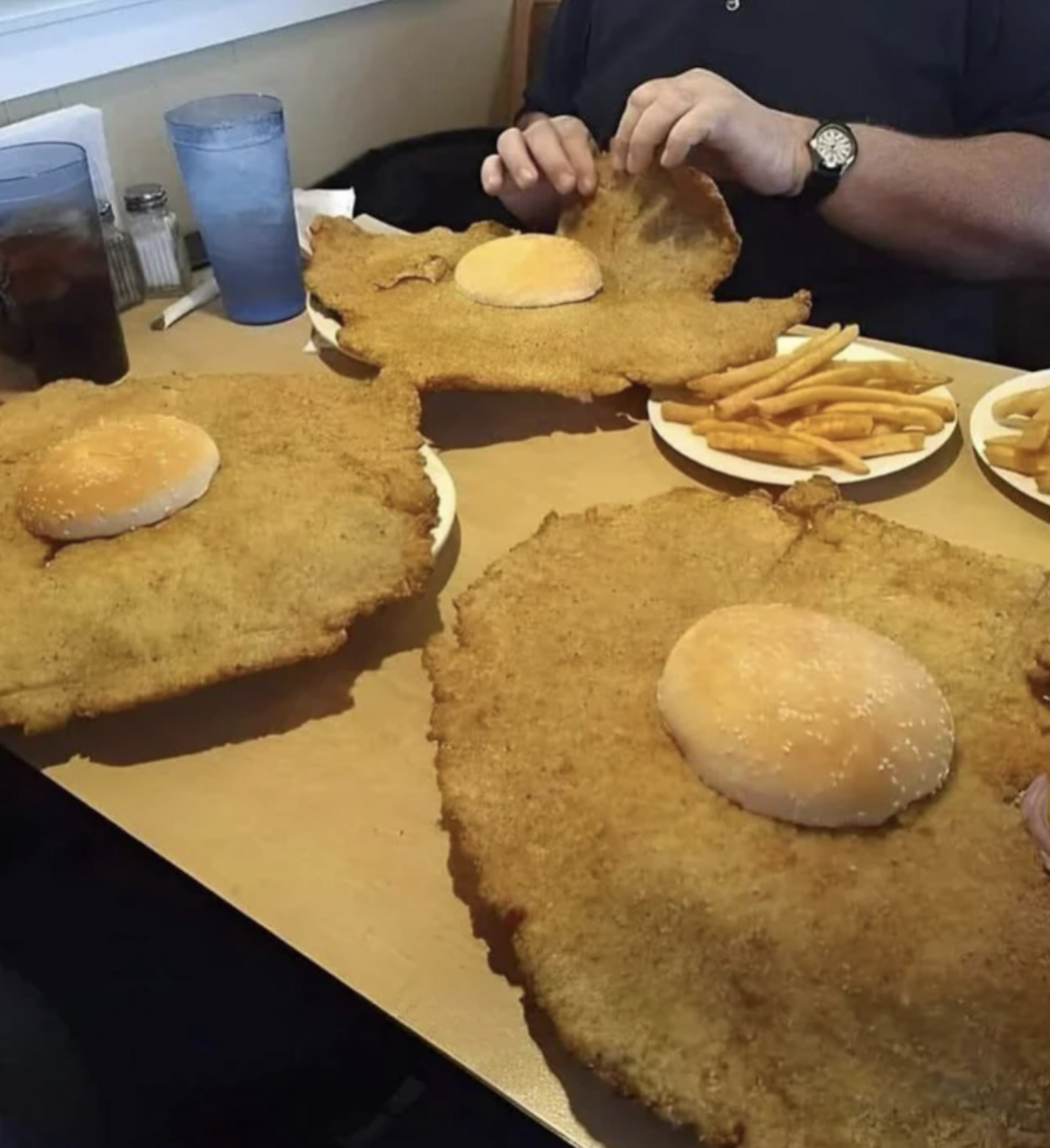 Three enormous schnitzels with small burger buns on top, served on plates with fries. Hands of a person are seen breaking a schnitzel. Glasses of drinks are in the background on the wooden table.