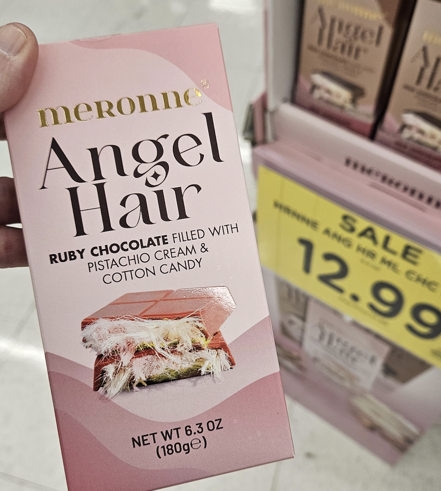 A person holds a box of Meronne Angel Hair Ruby Chocolate, filled with pistachio cream and cotton candy. A sale sign for $12.99 is visible on a shelf with more boxes in the background.