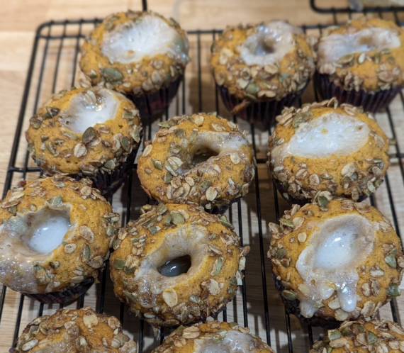 Twelve baked muffins with granola and seeds on top, each with a melted white icing spot in the center, are cooling on a black wire rack over a wooden surface.